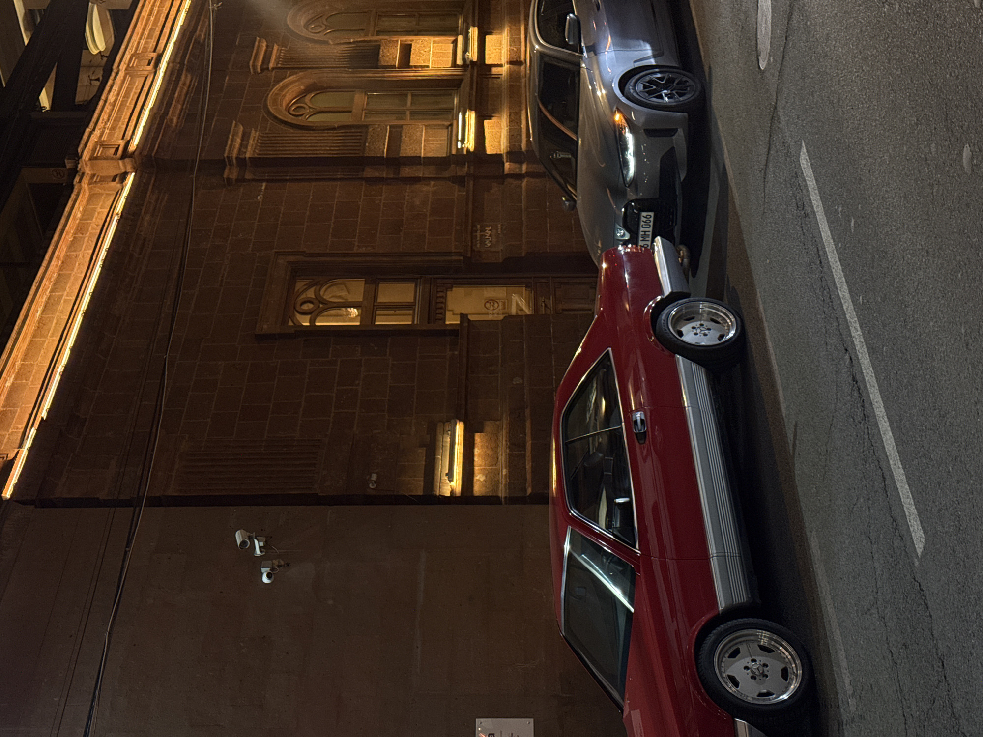 The image depicts a nighttime scene on a street in Yerevan, Armenia, featuring two parked vehicles: a red classic car and a newer model silver car. The cars are parked adjacent to a multi-story building constructed of dark masonry. The building's architecture includes arched windows, and the façade is partially illuminated by what appears to be architectural lighting. Surveillance cameras are mounted on the lower section of the building's wall. The street is paved and marked with a white lane divider. The street and adjacent building are dimly lit. The license plate visible on the newer car is '6 MH 066'.
