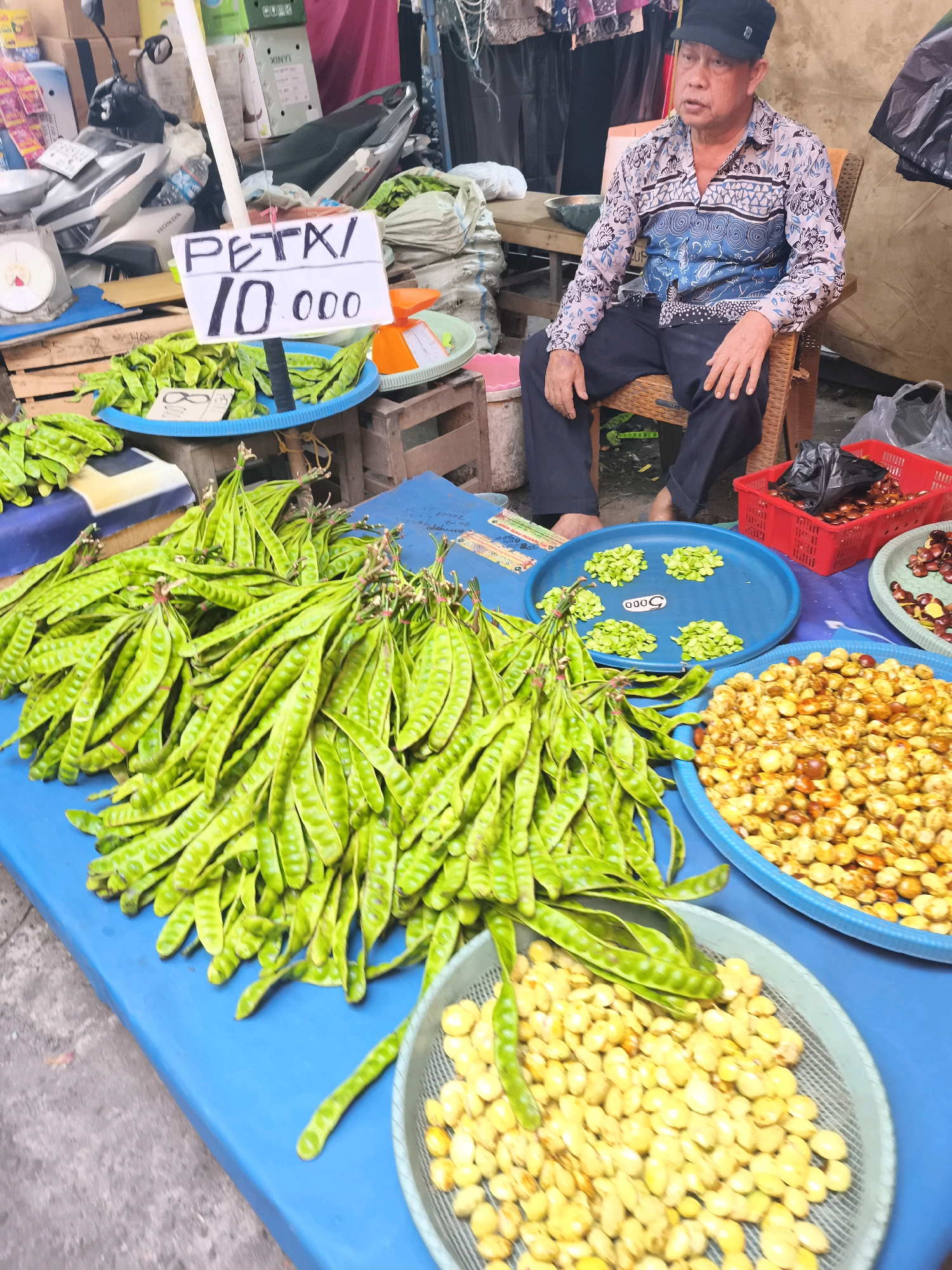 The image depicts a street market scene in Palembang, Indonesia, with a focus on produce. In the foreground, a vibrant blue table is laden with an abundance of fresh, green long beans, likely petai beans, a popular ingredient in Indonesian cuisine. These beans are bundled together and displayed prominently. To the right of the beans, a large blue plastic tray contains a pile of shelled, light yellow beans, possibly also petai, or a similar legume. Another plastic container to the right of that holds what appears to be a dried or roasted bean product with a brownish hue.
A white sign with black lettering, partially obscured by the beans, reads "PETAI/ 10.000," indicating the type of produce and its price in Indonesian Rupiah. In the background, a man wearing a black cap and a patterned shirt is seated on a woven chair, looking slightly to his left, possibly attending to customers or observing the market activity. Various other market elements are visible, including a motorbike, scales, baskets, and bags of produce, suggesting a bustling commercial environment. The overall atmosphere suggests a typical day at a local Indonesian market.