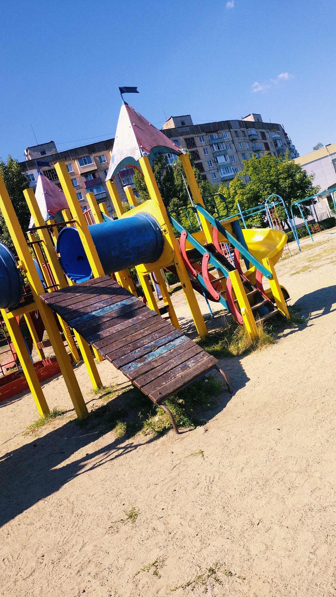This is a full-shot, eye-level, outdoor photograph of a brightly colored playground with a sand-covered ground. The playground structure is predominantly yellow and blue, with red and green accents. It features a slide, a tunnel, and stairs. A wooden ramp with weathered planks leads up to a section of the playground. In the background, a multi-story apartment building with many windows and a tiled roof is visible, along with green trees. The sky is clear and blue. The lighting suggests it is daytime, with shadows cast by the playground equipment onto the sand. The overall scene depicts a public recreational space on a sunny day.