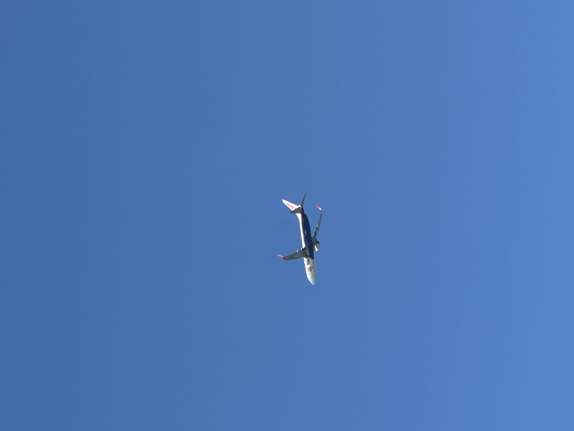 A commercial airplane with the logo of the airline visible on its fuselage is captured in mid-flight against a clear blue sky. The airplane is a single object in the frame, appearing in the right-center portion of the image. The sky is a uniform shade of blue, indicating a daytime scene with no clouds. The airplane is depicted from a side-angle, showing its wings, fuselage, engines, and tail fin. The location is unknown, and there are no people or other objects visible in the image.