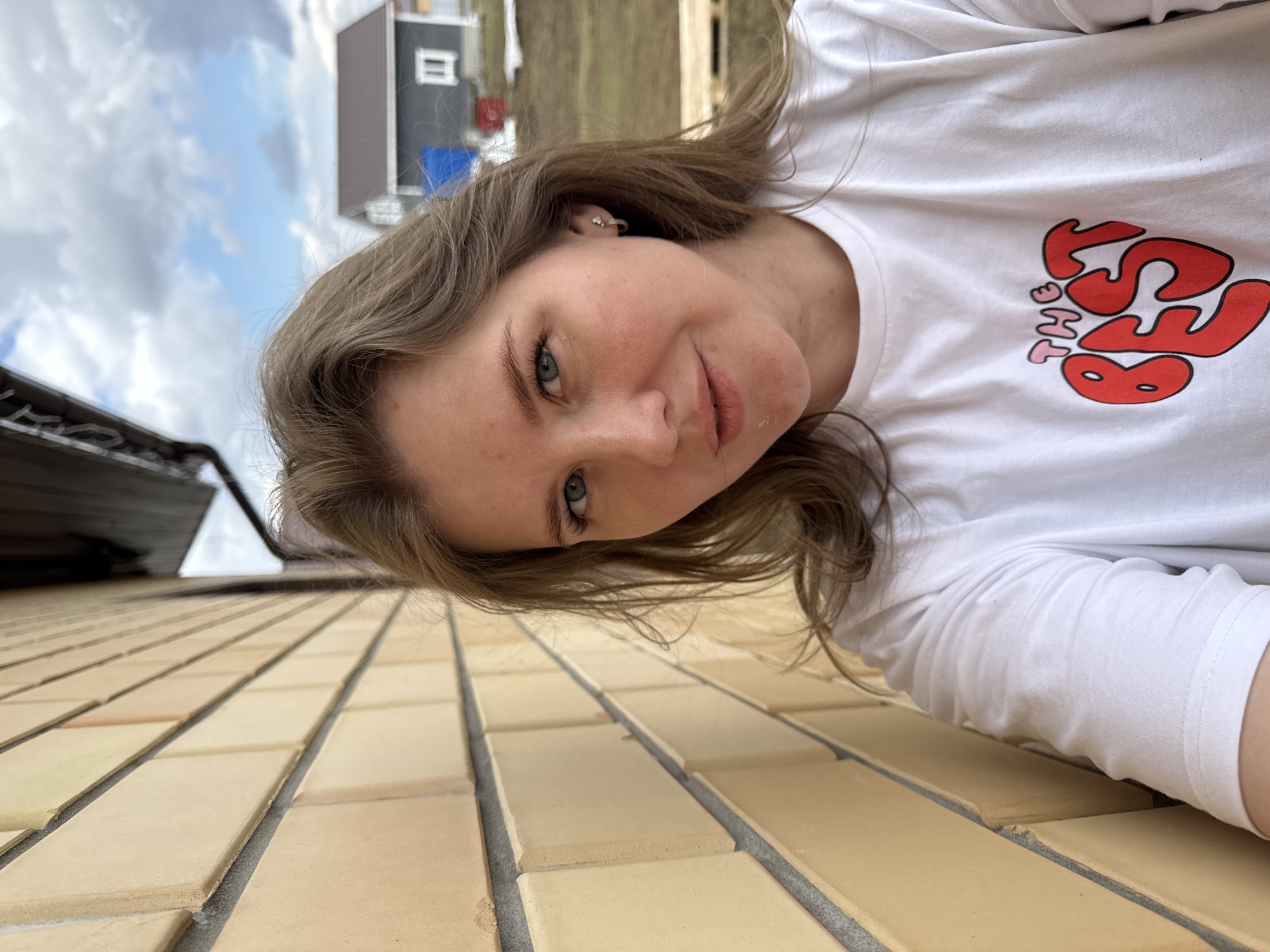 A young woman is captured in a close-up selfie, oriented diagonally within the frame. She has light brown hair, blue-green eyes, and fair skin, looking directly at the camera with a gentle smile. A small, simple earring is visible in her left ear. She is wearing a white long-sleeved t-shirt with stylized red and pink text on the chest that reads "THE BEST".
The image is taken from a low-angle perspective, with a light yellow/beige brick or tiled wall forming a prominent diagonal in the foreground and receding towards the upper left. The mortar lines between the tiles are gray.
In the background, under a partly cloudy sky with patches of blue, various outdoor elements are visible. To the left, a dark-colored building with a light-colored window is present, along with what appears to be a blue object near its base. A partial view of a roof edge or gutter is on the far left. Further to the right, a grassy or earthy hillside can be seen. The scene suggests an outdoor setting, possibly in a residential or construction area within Gornyi (Dvurechenskoe S/P), Russia, during daytime under overcast but bright conditions.