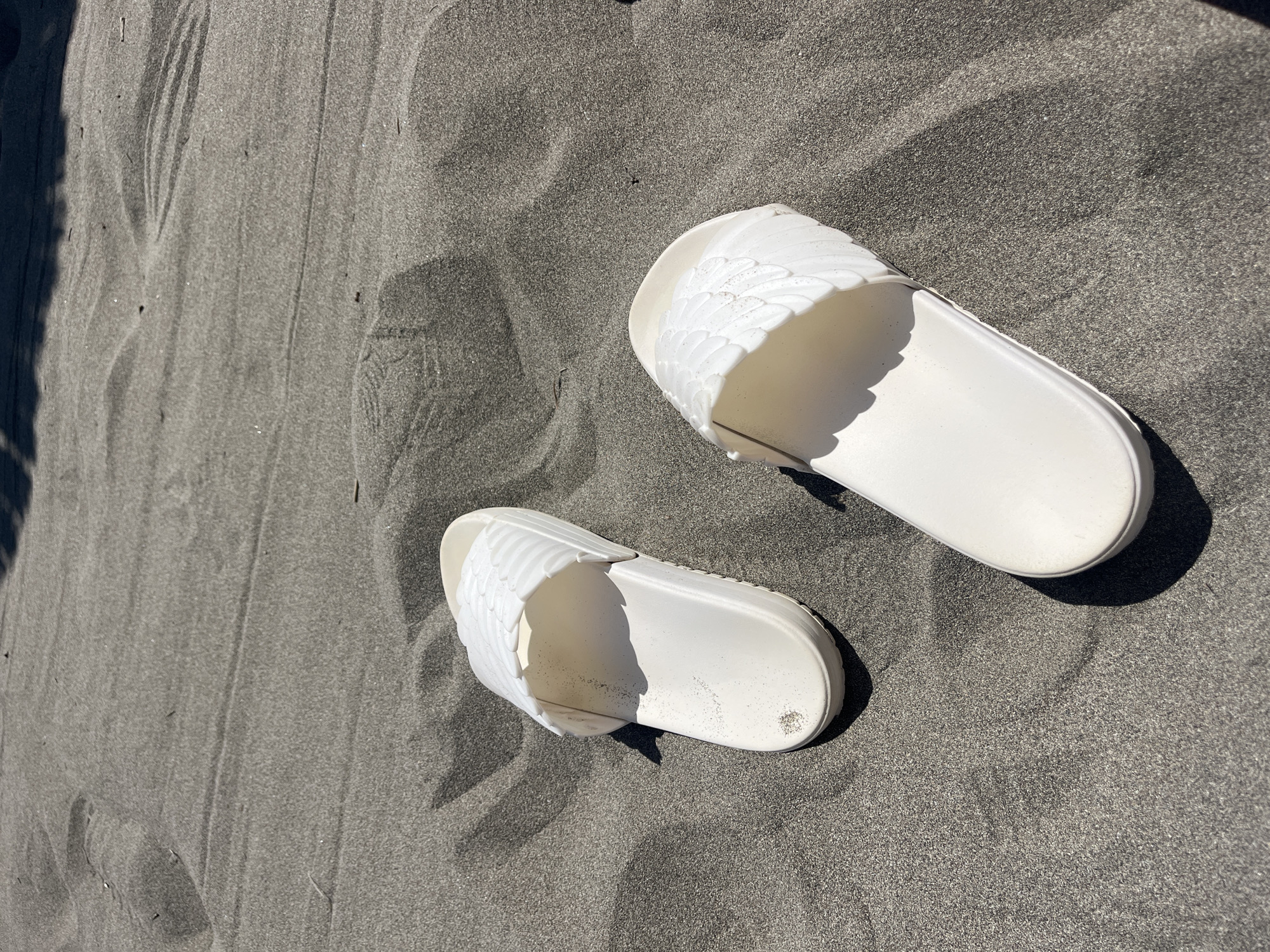 The image shows a pair of white, open-toed sandals with a decorative wing-like pattern on the upper part of each shoe resting on dark grey sand. The sandals are positioned slightly askew from each other, with one placed higher in the frame than the other. The sand appears to be fine-grained and covers the majority of the image. The sandals appear to be dusted with the sand. The location is Donji Štoj, Montenegro, given the presence of black sand, known to exist on the beaches near the city.