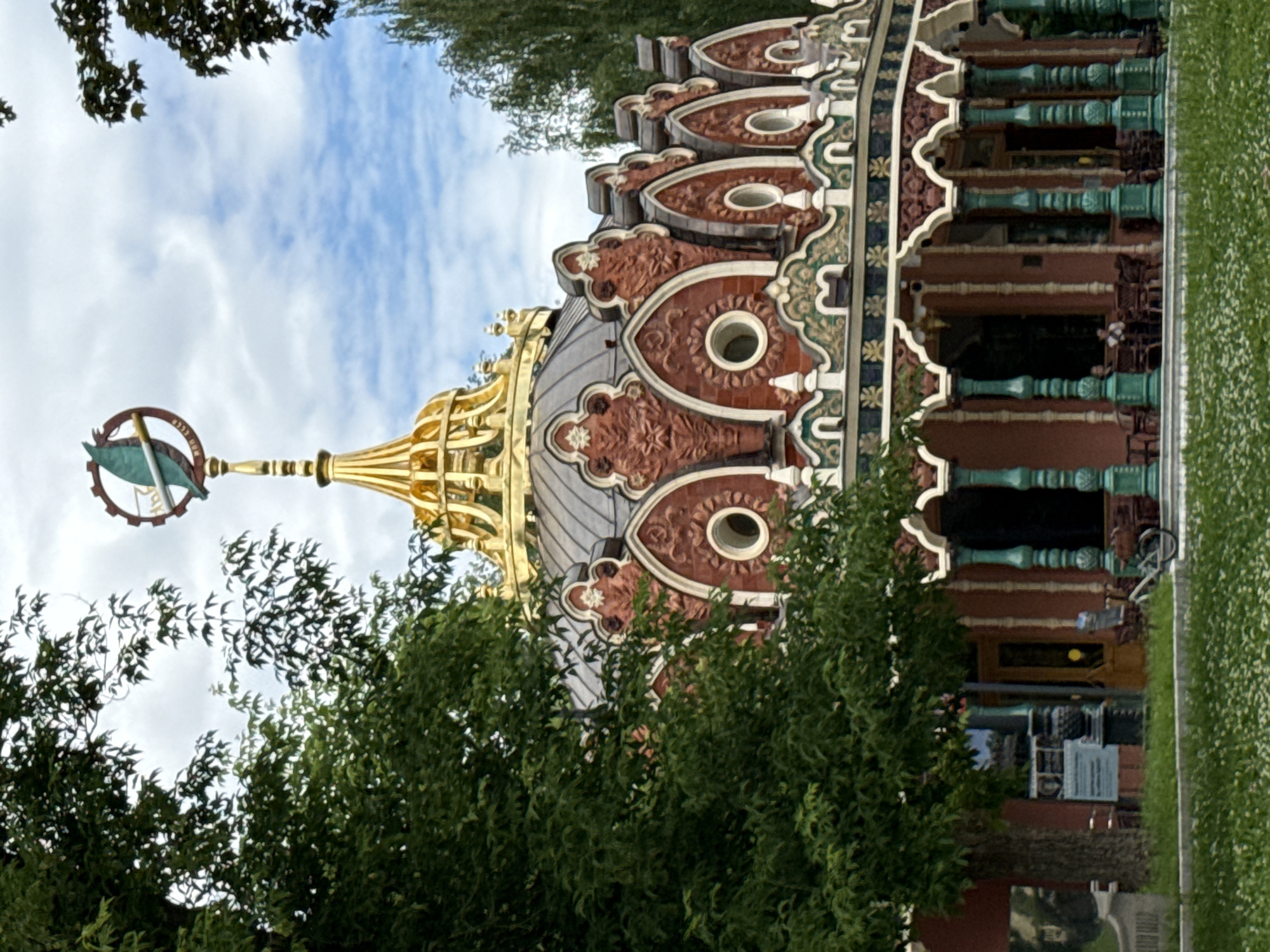 The image shows a detailed view of the exterior of the Miratorg Carousel building in Gorky Park, Moscow, Russia. The building features an ornate, multi-tiered roof with a golden dome and decorative red and gold accents. The facade is adorned with intricate carvings, arched windows, and colorful trim. Green, fluted columns support an open-air veranda, where several people are seated at tables. A bicycle is parked on the ground in front of the veranda. Trees with lush green foliage surround the building, and a cloudy sky forms the backdrop. The grounds in front of the building are covered in grass and small white flowers.