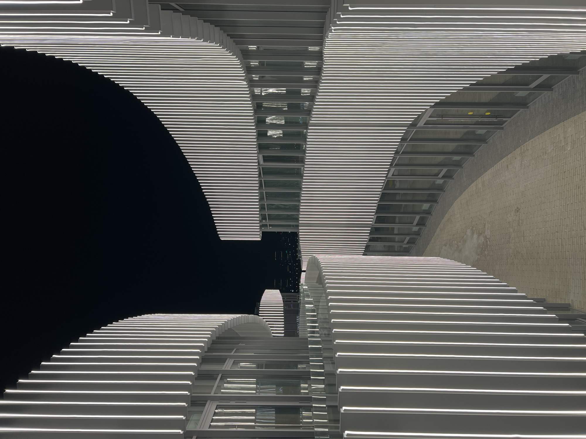 This image displays the illuminated exterior of the Kazan Expo International Exhibition Center in Kazan, Russia, at night. Multiple curvilinear building facades are visible, uniformly covered with closely spaced vertical strips of bright white light, creating a ribbed texture. The structures feature extensive glazing with numerous windows and doors on their lower levels, reflecting ambient light. A paved pedestrian walkway, constructed from light-colored rectangular pavers, winds between the curved architectural elements. No individuals or discernible objects are present within the immediate scene. Distant city lights are faintly visible against the dark, clear night sky in the background.