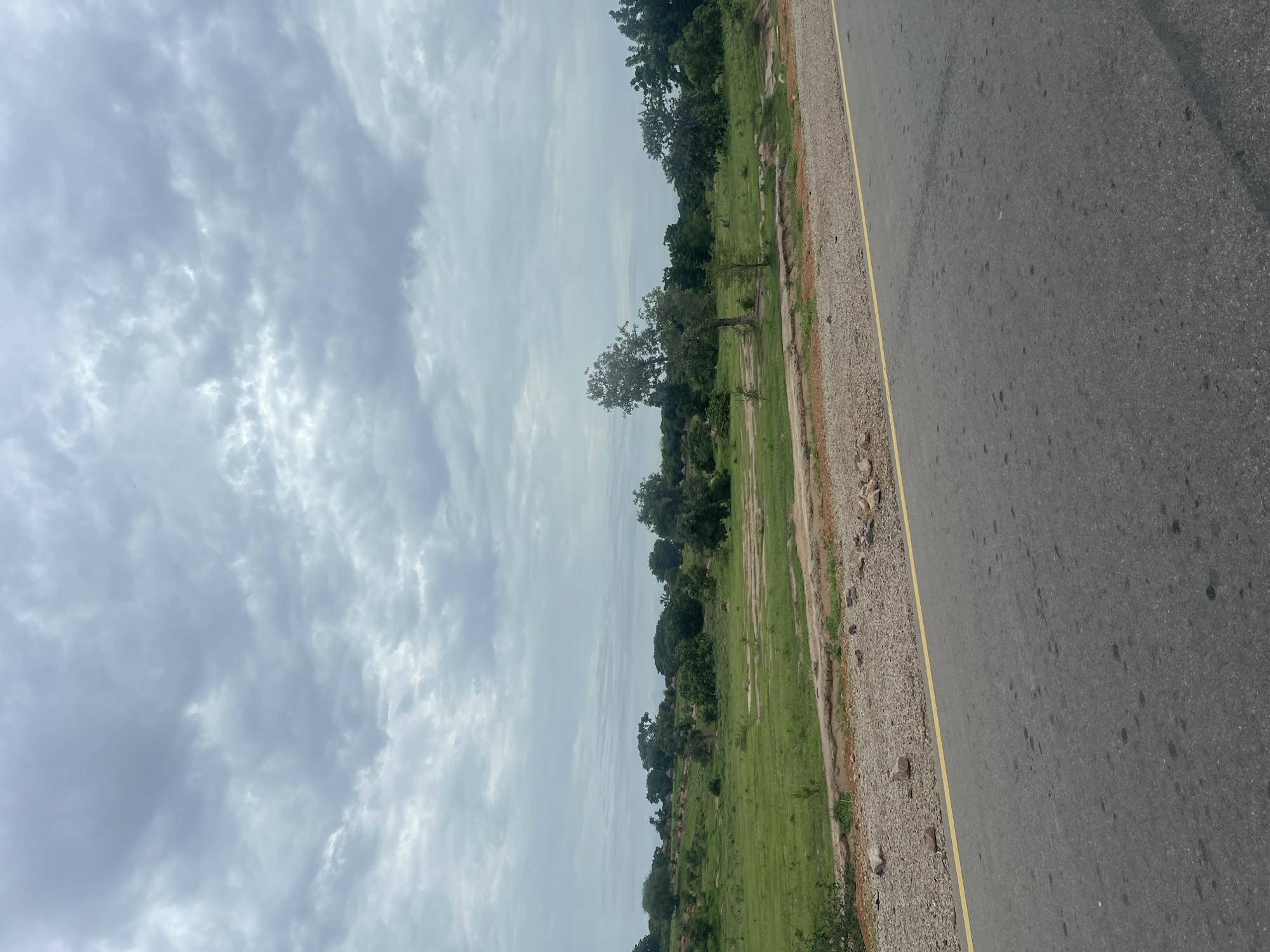 The image depicts a roadside scene in Minda, Nigeria, under a cloudy sky. The main subjects are the asphalt road with a yellow dividing line and a grassy area to its left. The road takes up the right side of the frame, appearing to be a two-lane highway. The grassy area features scattered trees and some sparse vegetation, with a sandy and rocky shoulder separating the grass from the road. In the distance, more trees and what appear to be small structures are visible, suggesting a rural or semi-rural setting. The sky is overcast with a mix of light and dark clouds, indicating it might be a cloudy day, possibly with a chance of rain. There is no visible activity or event taking place, and no people or distinct objects are discernible. The overall impression is one of a quiet, natural landscape alongside a transportation route. The time of day is not definitively clear but the lighting suggests it could be daytime. No text is visible in the image.
