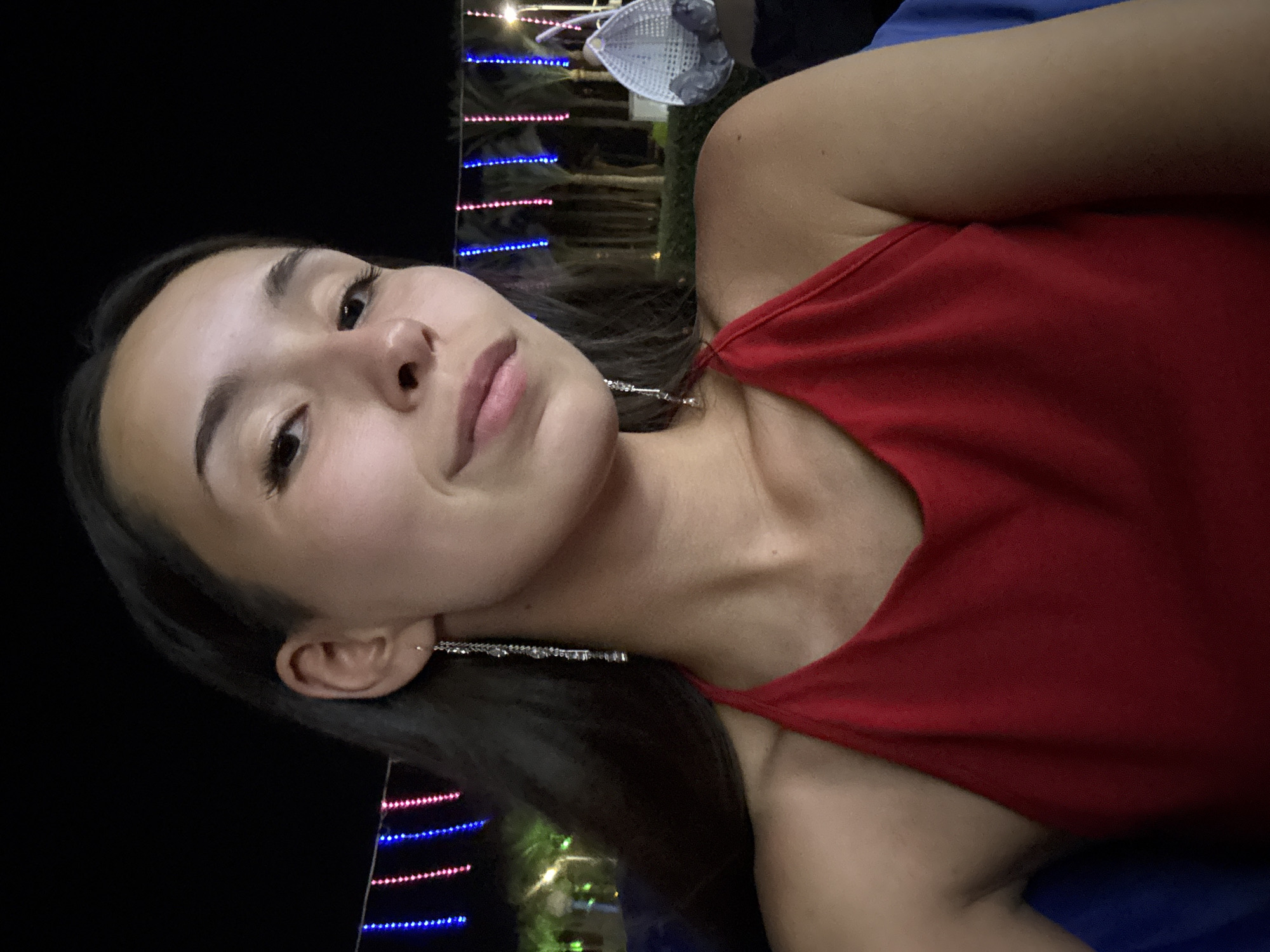 A close-up selfie of a young woman at night, wearing a red halter top and long, dangly earrings. She has dark hair and is looking towards the camera with a slight smile. In the background, there are palm trees illuminated by strings of blue and pink lights, and a white hanging chair. The setting appears to be an outdoor area, possibly a resort or entertainment venue, in an unknown location in Egypt.