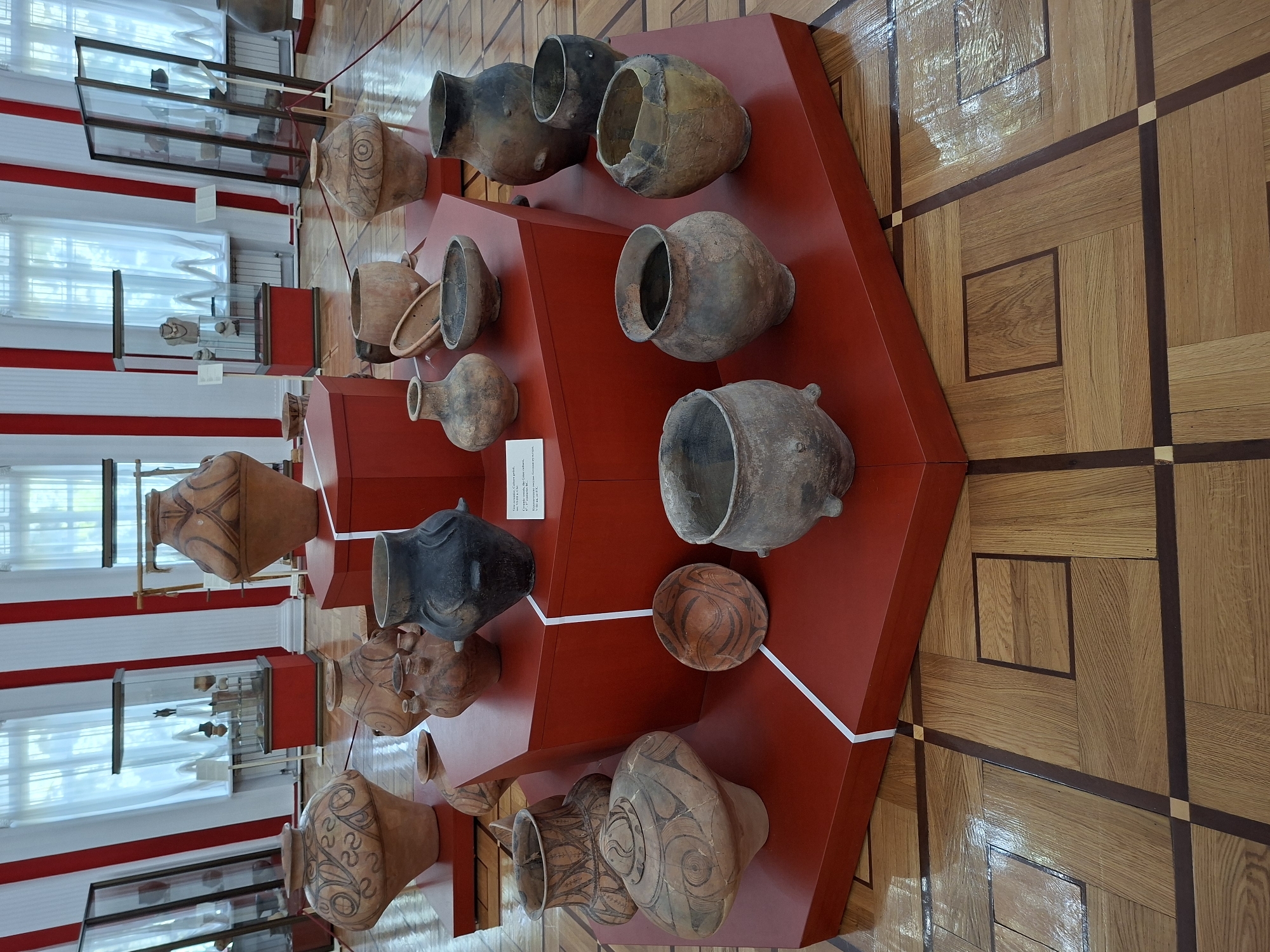 The image displays an indoor exhibition of ancient pottery, likely in a museum setting, featuring numerous earthenware vessels of varying sizes and designs. The pottery is arranged on a red, multi-tiered display platform in the foreground, with some additional pieces visible on the floor and in glass display cases in the background. The background also shows tall windows with white curtains, red and white striped vertical accents on the walls, and a polished wooden floor with a geometric parquet pattern.
Based on the provided crops and context, this exhibition is located at the National Museum of History of Moldova in Chișinău, Moldova. The museum is known to house artifacts from various historical periods of Moldova and surrounding regions. The specific exhibition appears to be focused on ancient pottery, possibly from Scythian or Sarmatian cultures given the styles of some of the vessels. There are no people visible in the image. The lighting suggests a well-lit indoor environment typical of a museum display. The scene is static, with objects placed for viewing and preservation.