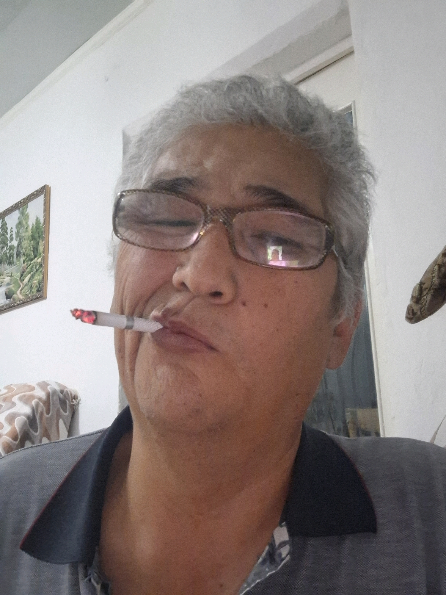 The image is a close-up, selfie-style photograph of an individual with graying hair and glasses, holding a lit cigarette in their mouth. The person is wearing a dark collared shirt. The background appears to be an indoor setting with a white wall and a framed picture on the left. A glimpse of what looks like a doorway or window is visible on the right. There is no information or visual cue in the image that would suggest the location is Almaty, Kazakhstan.