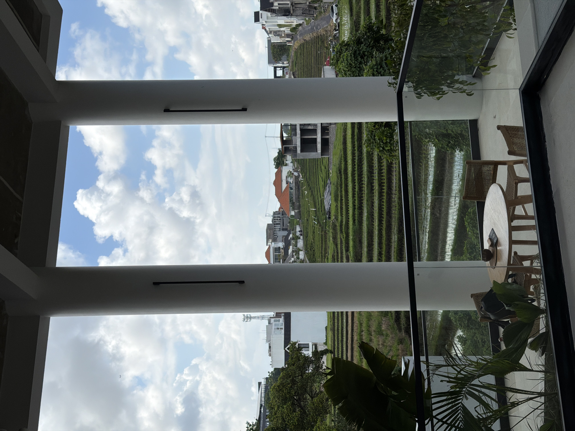 The image shows a view from what appears to be an indoor seating area overlooking rice terraces. The architecture consists of white pillars and a glass window or partition with a black frame. Through the pillars, the rice terraces are visible, and the sky above contains a mix of blue and white clouds. Beyond the terraces, there are buildings, giving the impression of a rural or suburban setting. Inside, a round table and chairs are seen. Green plants are present near the glass. This scene appears to be captured in Mengwi, Indonesia, based on the prevalence of rice terraces in the area. There are no visible people or actions directly captured in the image.