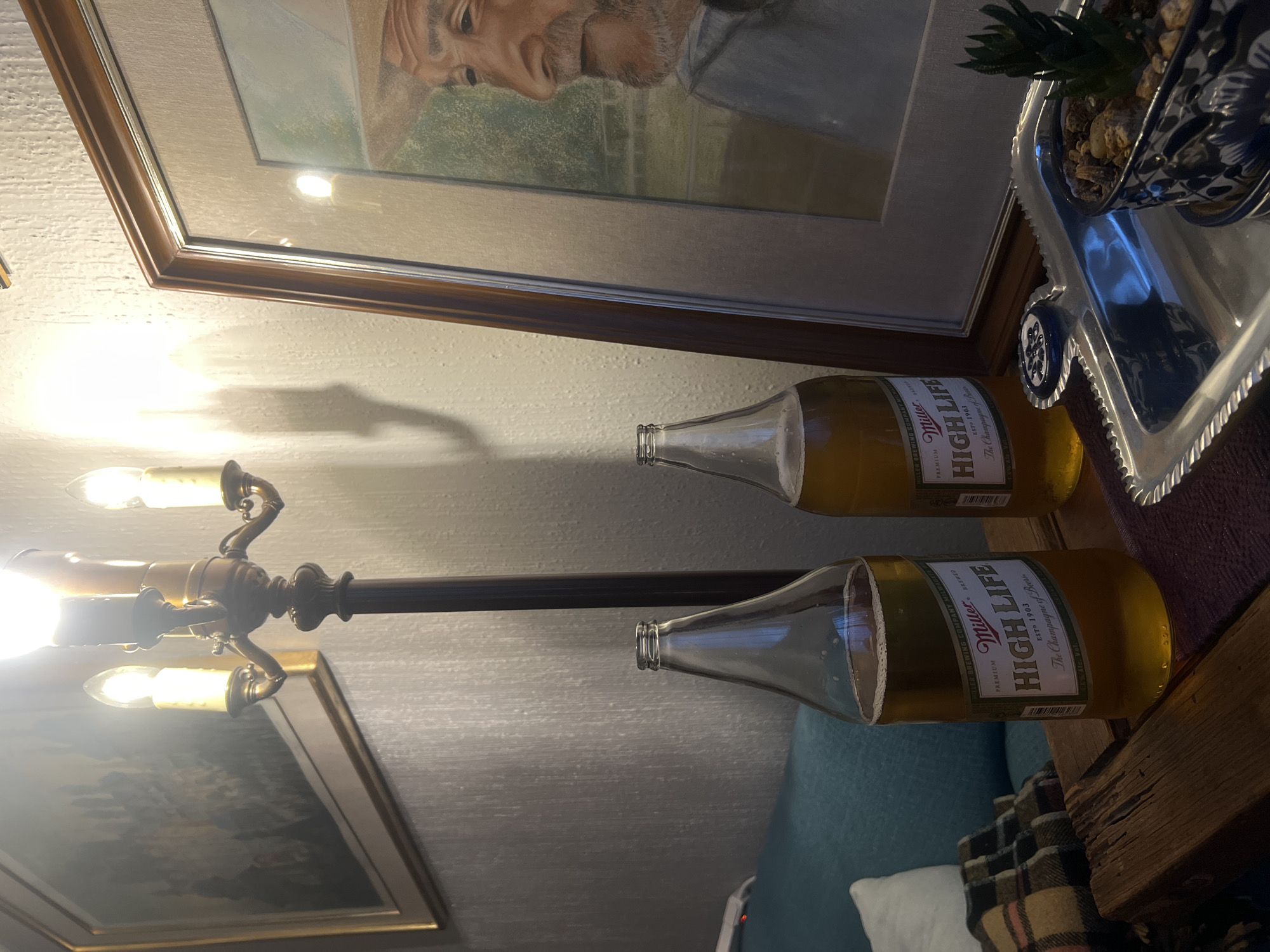 The image shows a cluttered indoor setting. Prominently displayed are two bottles of Miller High Life beer. Above them is a framed painting depicting a man, and to the right, a silver tray holding a potted succulent. Below the beer bottles and slightly to the left, a chandelier is visible, along with another framed picture. In the background, a glimpse of a blue couch with white pillows and a plaid blanket is discernible. The image does not contain any identifying information to determine if it was taken in Mexico City, Mexico.
