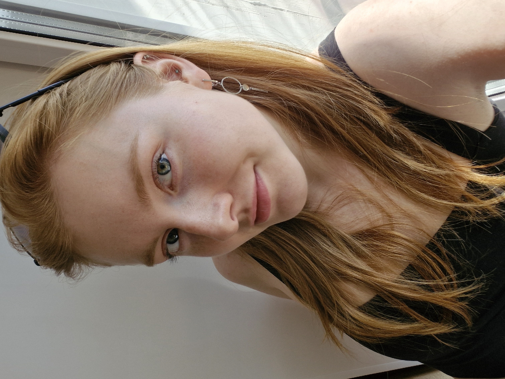 A young, fair-skinned woman with red hair is pictured indoors, likely near a window. She has green eyes and is wearing a black top. A silver earring is visible in her left ear. Sunglasses are perched on her head. The background appears to be a white or light-colored surface. There is no indication of the location being Yekaterinburg, Russia.