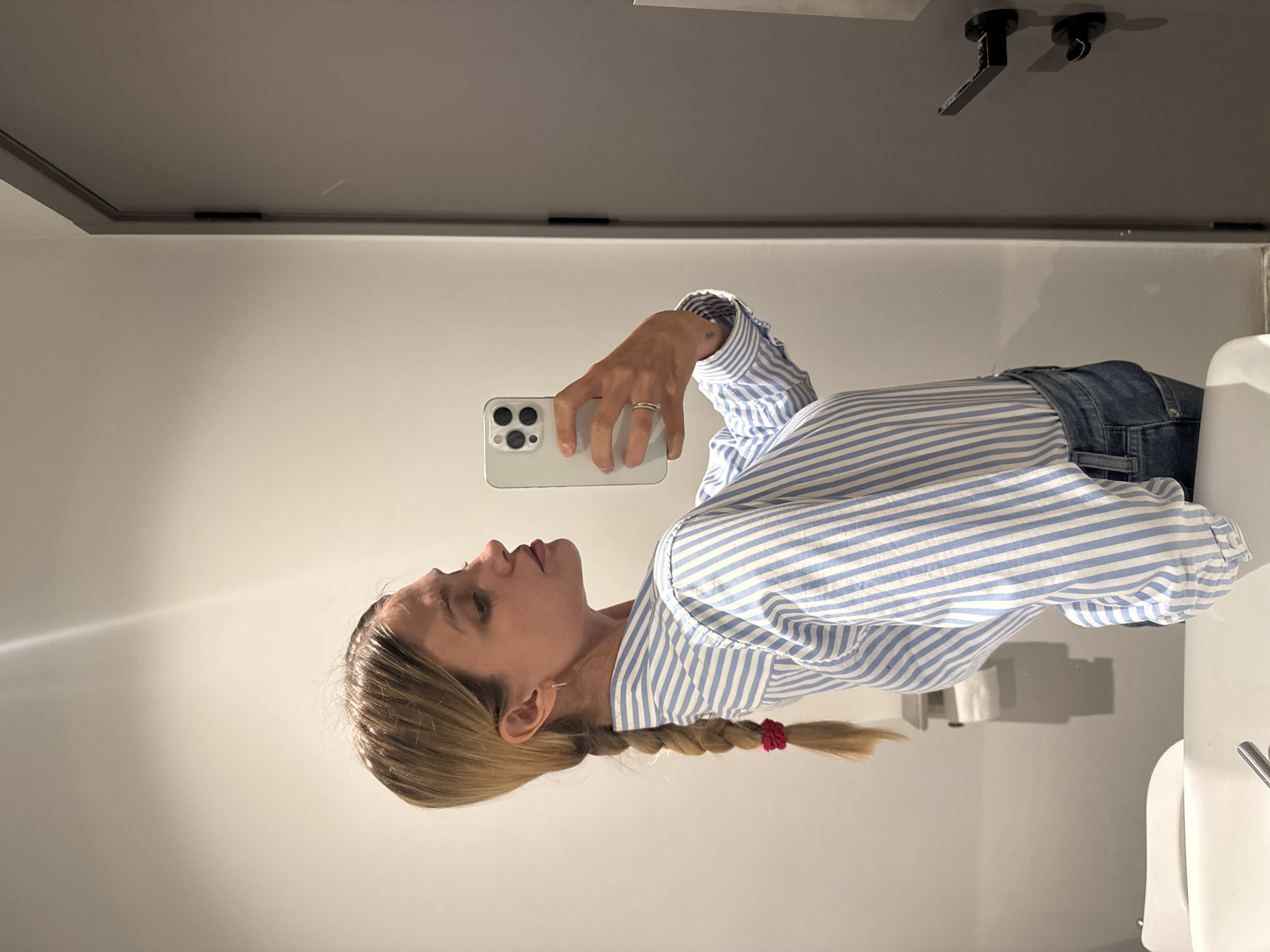 A woman with blonde hair, styled in a side braid secured with a red hair tie, is depicted taking a mirror selfie. The image is rotated 90 degrees counter-clockwise, showing her from the chest up, with her head tilted slightly upwards and to the right. She is holding a light-colored smartphone (an iPhone with three camera lenses visible) in her left hand, which has a ring on one finger. A small tattoo is visible on her left wrist. She is wearing a blue and white striped long-sleeved button-down shirt and blue jeans.
The setting appears to be an indoor bathroom, likely a public restroom or hotel bathroom, given the clean, modern aesthetic and fixtures. The background, reflected in the mirror, shows a plain light-colored wall and a darker grey ceiling with a recessed border. Two black, minimalist fixtures are mounted on the ceiling, possibly lights or handles for a door above the mirror line. Part of a white sink is visible on the right, and further back, a white toilet paper holder with toilet paper confirms the bathroom environment. The lighting is bright and even, indicating an indoor setting. No specific time of day or emotions are discernible beyond a neutral expression focused on taking the photo. No visible text is present in the image. The location context provided is Tbilisi, Georgia.