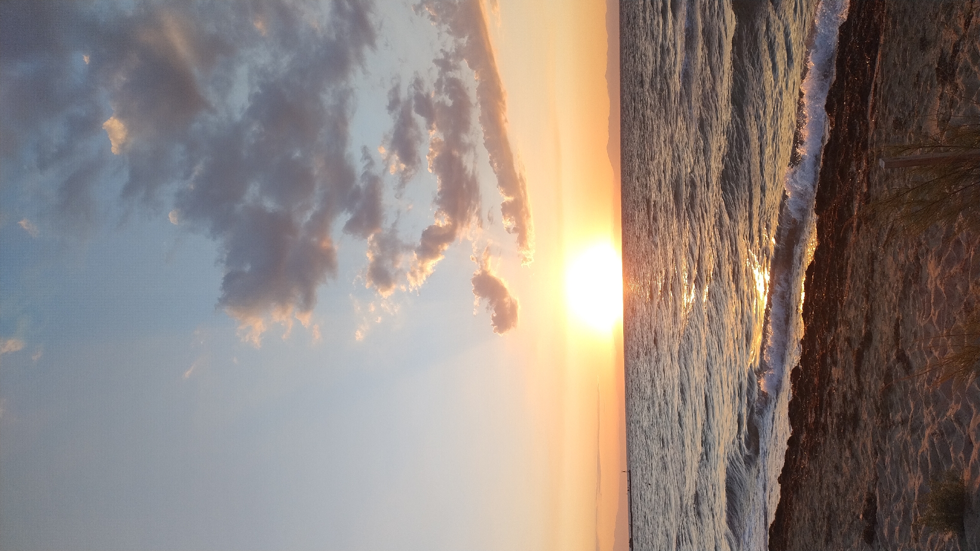 This photograph captures a serene sunset over the Aegean Sea, viewed from the beach in Neoi Epivates, Greece. The sun, a brilliant orb, is low on the horizon, casting a warm golden glow across the water and illuminating the sky with hues of orange, pink, and pale blue. Wispy clouds, tinged with the colors of sunset, drift across the sky. Gentle waves lap at the sandy shore, where a line of dark seaweed is visible. In the distance, faint outlines of land or mountains can be seen. A small pier or structure with a few indistinct figures is present on the left side of the frame. The sand on the beach appears light in color, with some sparse vegetation in the foreground. The overall atmosphere is tranquil and picturesque.