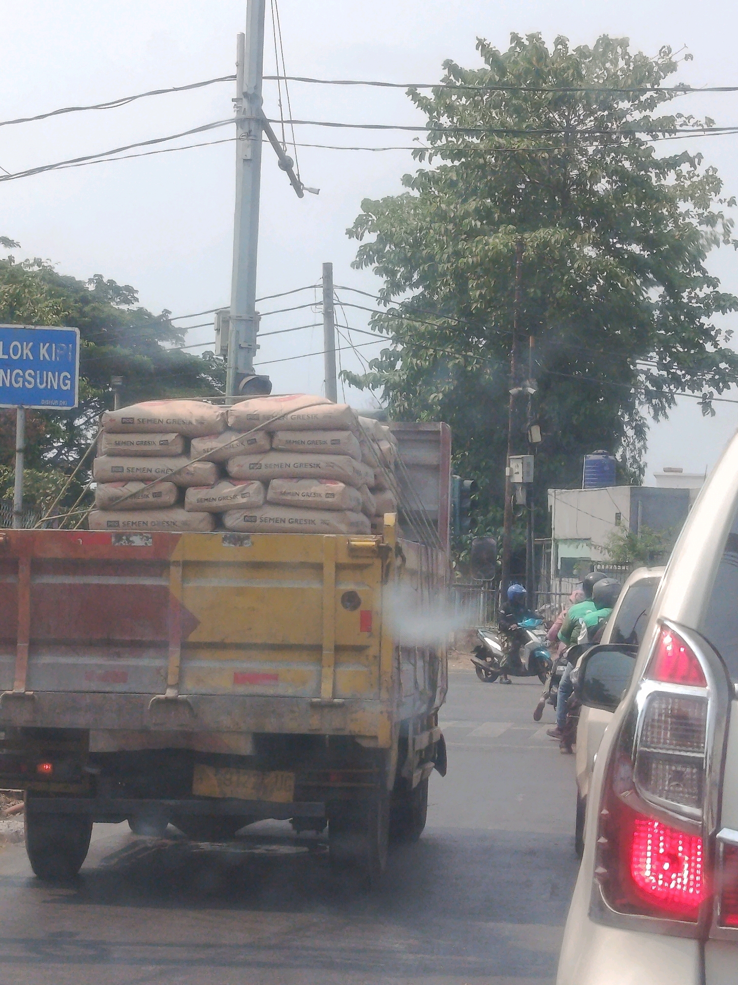 The image shows a street scene in East Jakarta, Indonesia. A truck laden with sacks of cement, labeled "SEMEN GRESIK", is in the foreground. The truck is yellow and red. Behind the truck, a white car's taillight is visible. Several motorcycles are in traffic behind the truck, with riders wearing helmets and casual clothing. Some are wearing blue, green, and black helmets. Trees are present on the side of the road, and utility poles with wires are visible overhead. A blue sign with white text, partially obscured, is also visible on the left. The sky is overcast.
