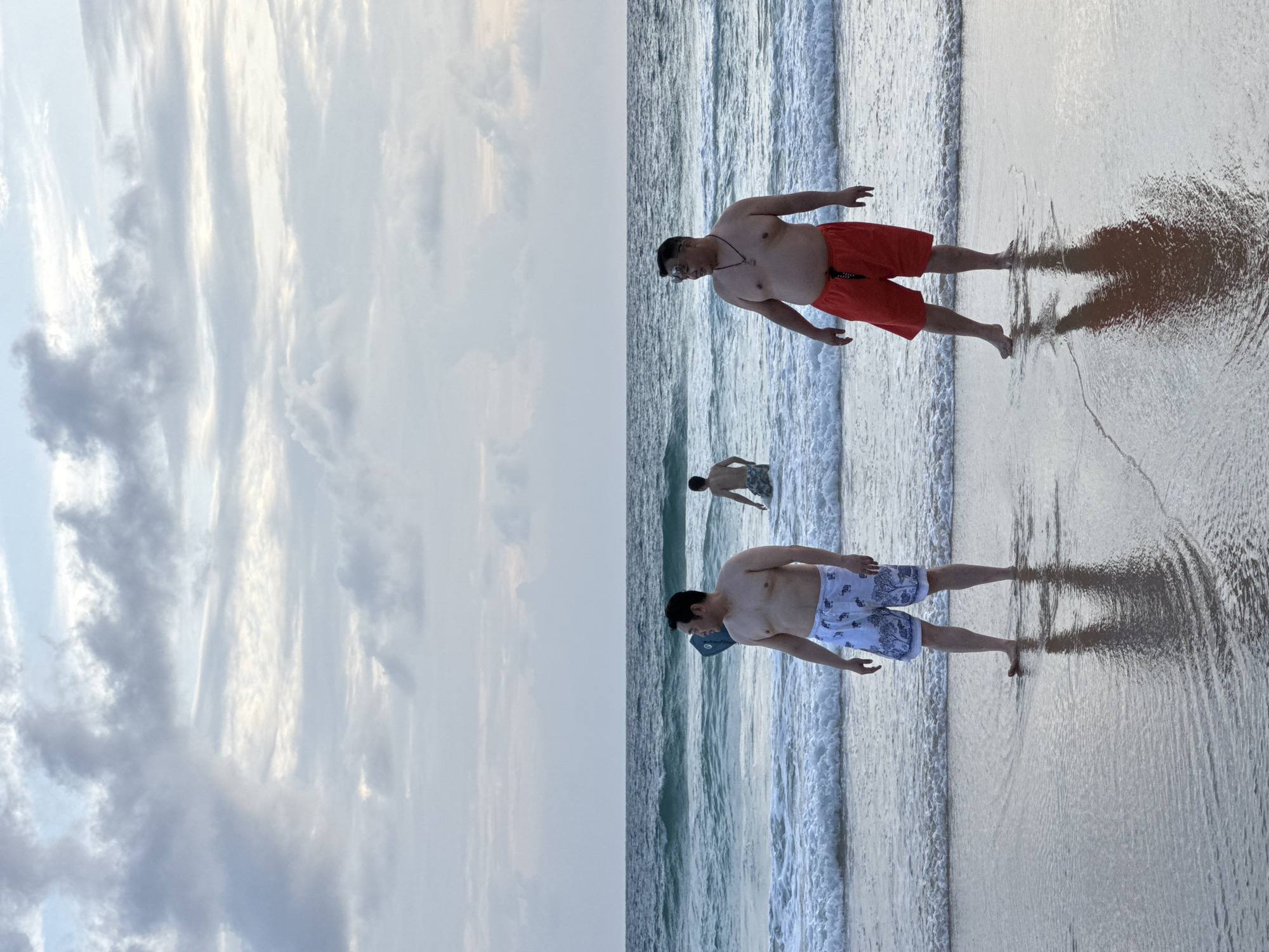 In Karon, Thailand, two men are standing in the shallow surf of the ocean. The man on the left is wearing white swim trunks with a blue elephant print. The man on the right is wearing red swim trunks. Both men are shirtless. In the background, another person is in the water further out. The sky is overcast with clouds. The waves are breaking on the shore, and the water is a deep blue-green. The sand is wet and reflective.
