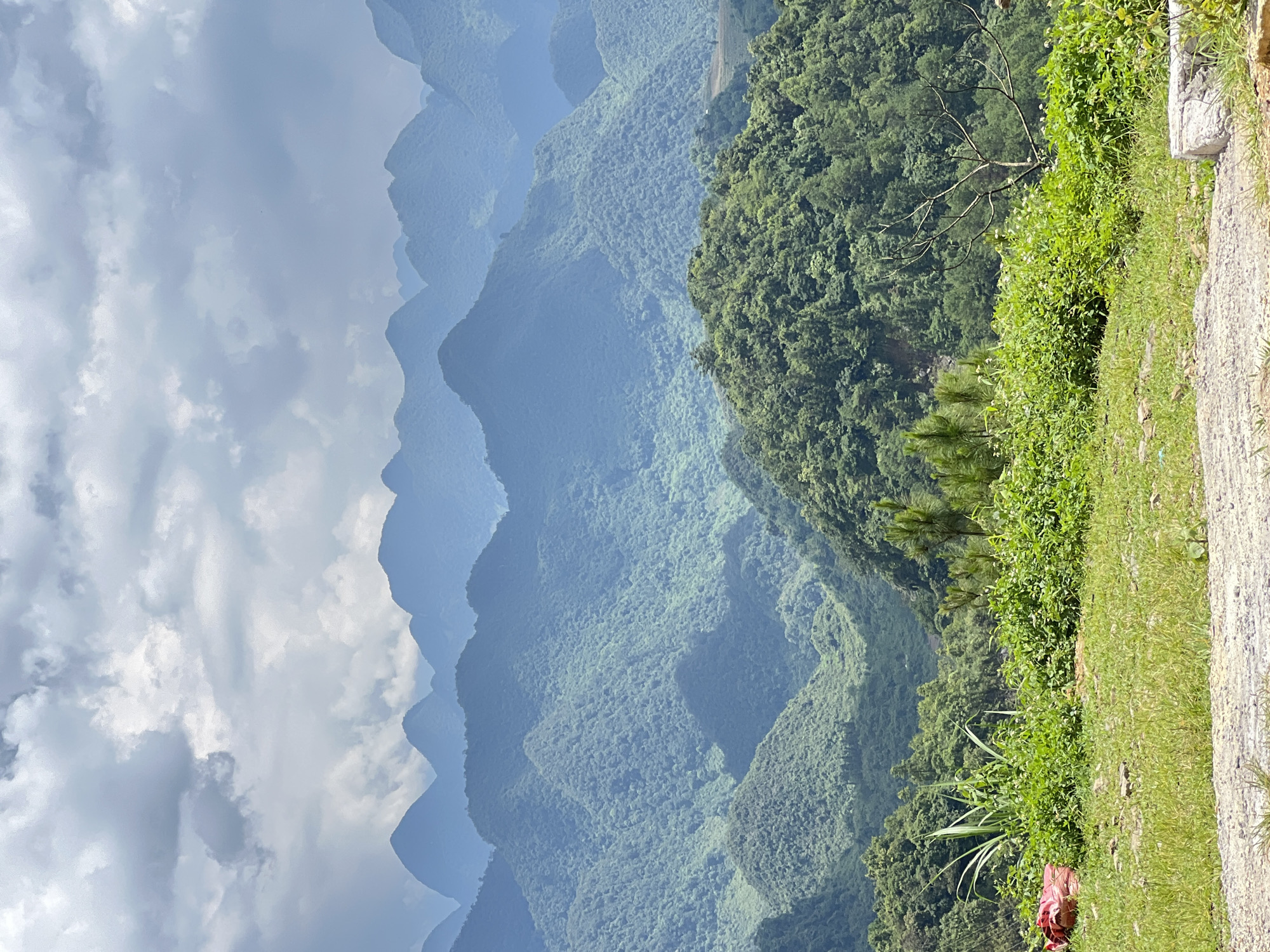 The image captures a panoramic view of verdant, rolling mountains under a cloudy sky. The foreground features a grassy area with scattered rocks and a patch of dirt. To the left, a red bundle is visible amidst lush green foliage. The middle ground is dominated by densely forested hills, with the trees creating a textured, emerald carpet. In the distance, a series of majestic mountains rise, their peaks fading into a hazy blue, suggesting depth and scale. The overall impression is one of natural grandeur and serenity. This vista is characteristic of the breathtaking landscapes found in Quản Bạ Commune, Vietnam, known for its dramatic karst topography and lush vegetation.