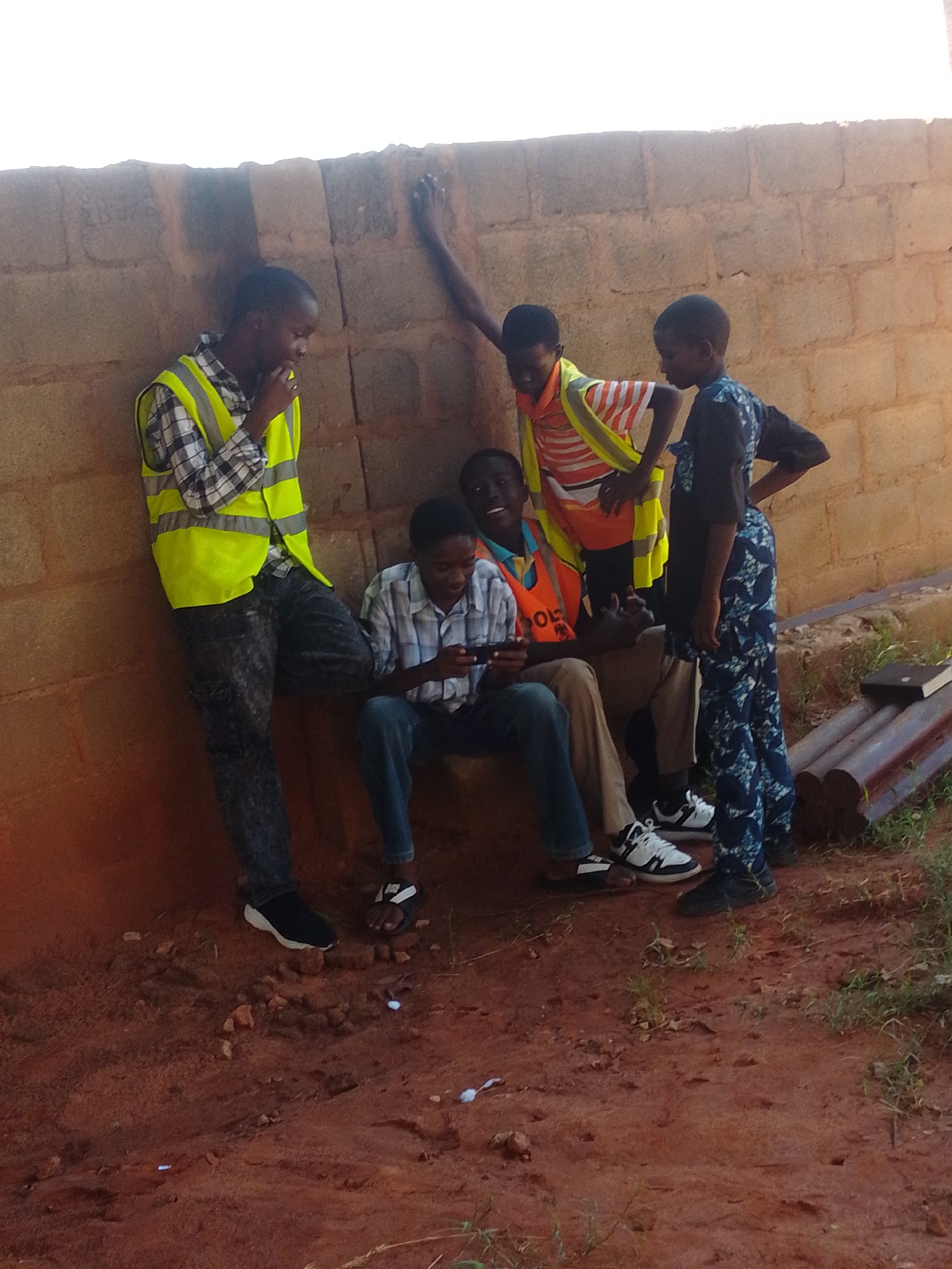 In Tumfure, Nigeria, four young individuals are gathered near a textured, reddish-brown brick wall. Two stand while two sit. The standing individuals are on the left and right. The person on the far left wears a checkered shirt and a neon yellow safety vest. They lean against the wall, with their right hand near their face, looking down. To their right, another person stands wearing a striped orange and white shirt with a neon yellow safety vest, and a bright orange vest over it. They are looking down and to the left, with their left hand on their hip.
Seated on the ground are two other individuals. The one on the left is wearing a light-colored checkered shirt and blue jeans, and is holding a smartphone, looking at it. Next to them, to the right and slightly behind, is another person, partially obscured, wearing a dark patterned garment. They are looking forward and to the right.
The ground is a mixture of reddish-brown soil and scattered small rocks. To the right of the seated individuals, there are several dark, cylindrical objects, possibly pipes, and a black book resting on them. Green vegetation is visible in the background, along the base of the wall and near the pipes. The lighting suggests it is daytime, with shadows cast on the ground.
