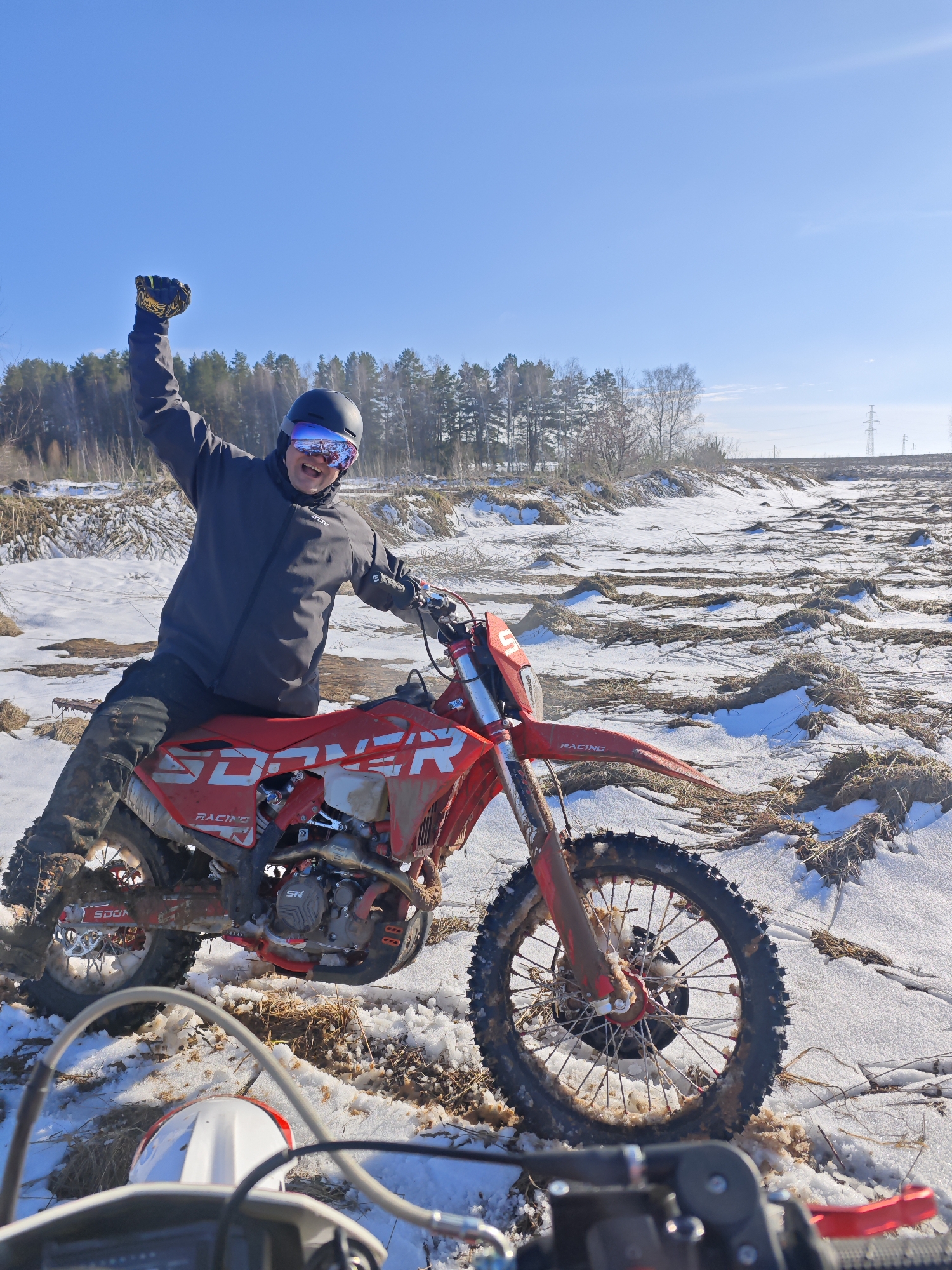 A man is sitting on a red dirt bike in a snowy field. He is wearing a black jacket, black helmet, and goggles. He has his right arm raised in the air. The dirt bike has the word "SOONER" on the side and the number 5 on the front. The field is covered in snow and dead grass. There are trees in the background. The location is Прикамский, Russia.