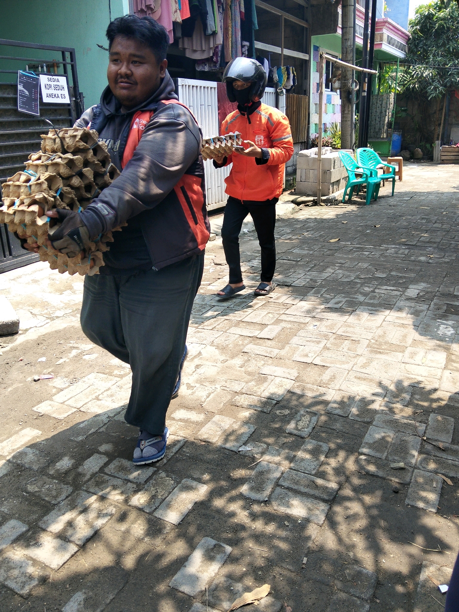 In the city of Rajegmulya, Indonesia, two individuals are depicted on a cobblestone pathway. The foreground features a man with dark hair and a mustache, wearing a black jacket with orange accents over a dark shirt and grey pants. He is carrying a large stack of brown cardboard egg cartons. To his right and slightly behind, another person, wearing a black helmet and a black face mask, is clad in an orange jacket and black pants, also holding a stack of egg cartons. The environment suggests an outdoor setting with buildings visible in the background, including one with a green facade and another with pink and white accents. There is also some greenery and what appears to be a blue plastic chair to the right of the second individual. The pathway is partially shaded by tree leaves.
