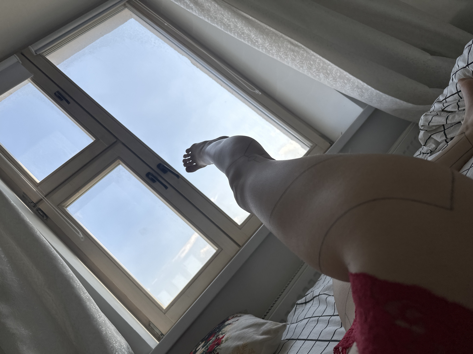 A low-angle, indoor shot shows a person's leg and foot in the foreground, positioned in front of a window. The leg is smooth and lightly toned, with subtle lines or markings visible on the skin. The foot is extended towards the upper left, displaying the toes. The person appears to be wearing red underwear.
The window is a multi-paned, rectangular structure, allowing a view of a bright, clear sky with some clouds. The window frame is light-colored. Curtains are visible along the edges of the window, adding a sense of softness to the scene.
Beneath the leg, there is a bed with white and black striped bedding. A pillow with a floral pattern is also visible.
Based on the available information, it is impossible to verify the location to be Khimki, Russia. The scene appears to be a casual, indoor setting, possibly a bedroom.
