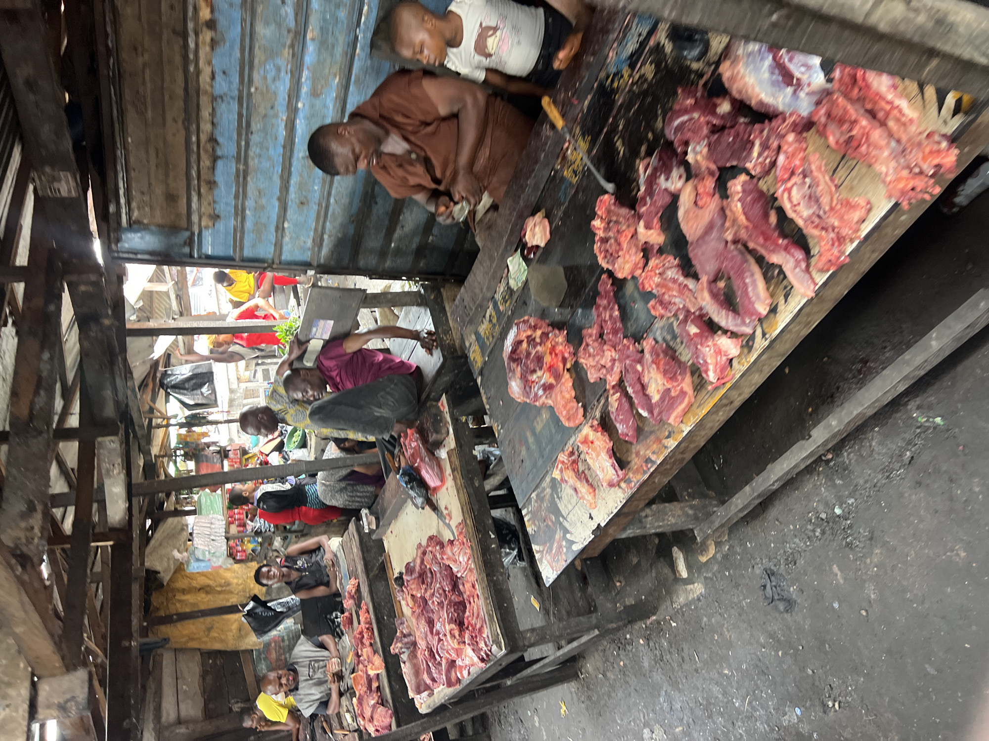 The image depicts an outdoor meat market in Nkpor, Nigeria. There are several vendors selling raw meat laid out on wooden tables. Customers and passersby are visible in the background. The vendors are cutting and preparing the meat. Overhead, a patchwork of wooden and metal structures provides partial shelter. The ground appears dirty and worn. The overall impression is a bustling, informal market setting.