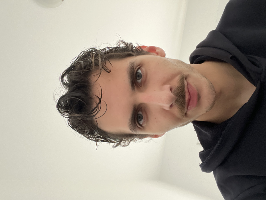 The image presents a head-and-shoulders portrait of an adult male, taken indoors. The subject has dark, wavy hair that appears wet or styled, dark eyebrows, and blue-green eyes. He features a dark, well-groomed mustache and light stubble on his chin and jawline. His lips are closed, forming a neutral to slightly curved expression. The male is wearing a dark-colored, likely black, hooded sweatshirt with visible fraying along the collar edges. He is looking directly at the camera. The background consists of a plain, light-colored wall. A circular, light-colored object, potentially a light fixture, is partially visible in the upper right corner of the background. The lighting is even and appears to originate from an internal source. No specific elements within the image identify the location as Bečići, Montenegro.