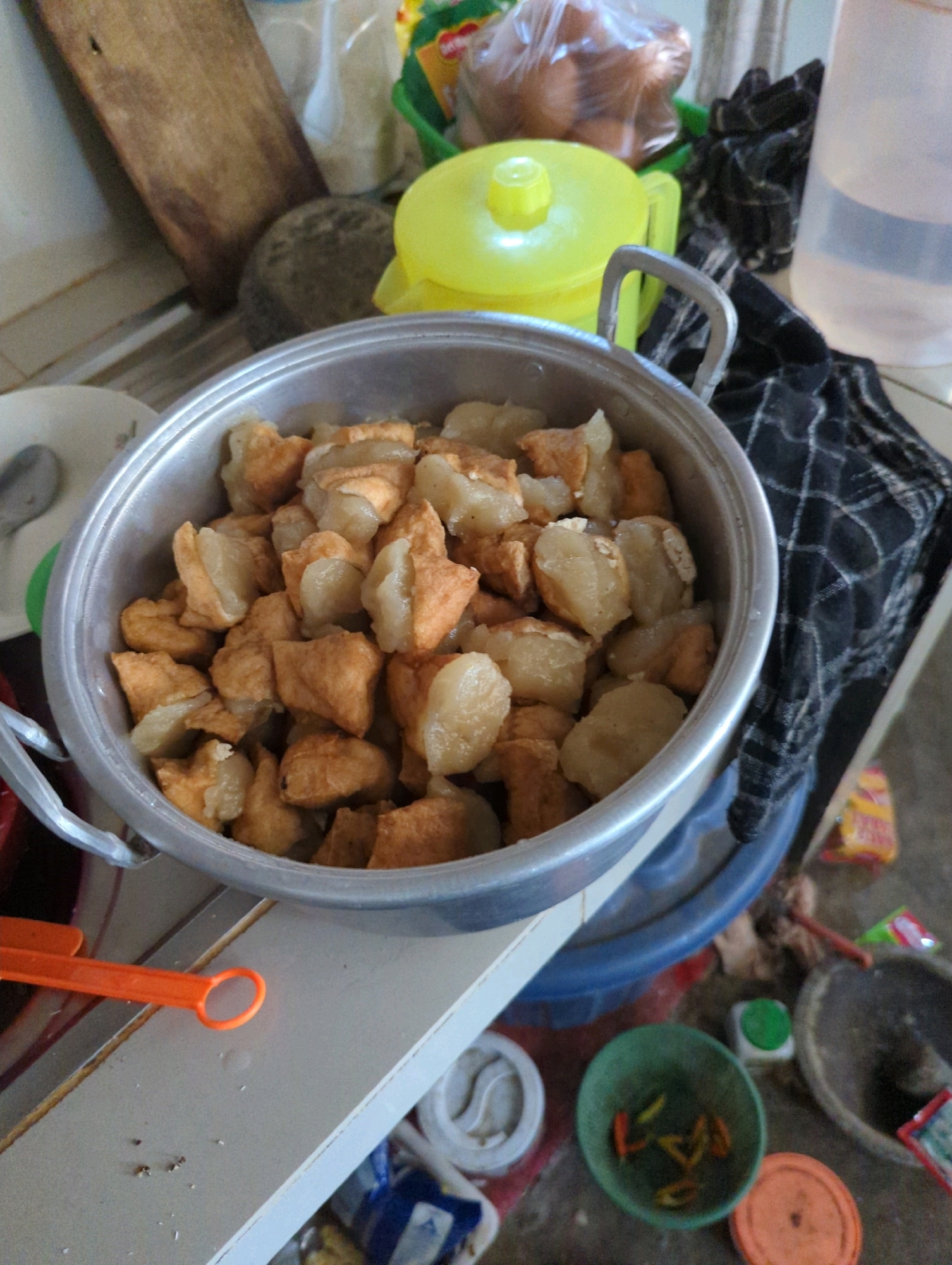 In a brightly lit kitchen area, likely in Senggrong, Indonesia, a large metal pot overflowing with fried tofu and dumplings takes center stage. The pot, with its two silver handles, rests on a blue plastic basin. Surrounding the pot, various kitchen items are scattered: a weathered wooden cutting board, a grey mortar and pestle, a bright yellow lid to a container, and a black and white checkered cloth. In the foreground, an orange plastic tool with a circular handle lies on a white surface, next to some scattered crumbs. In the background, a clear plastic bottle filled with water and a green basket containing chili peppers are visible, along with other unidentifiable objects and packaging. The overall impression is one of a domestic kitchen scene, possibly mid-meal preparation or serving.