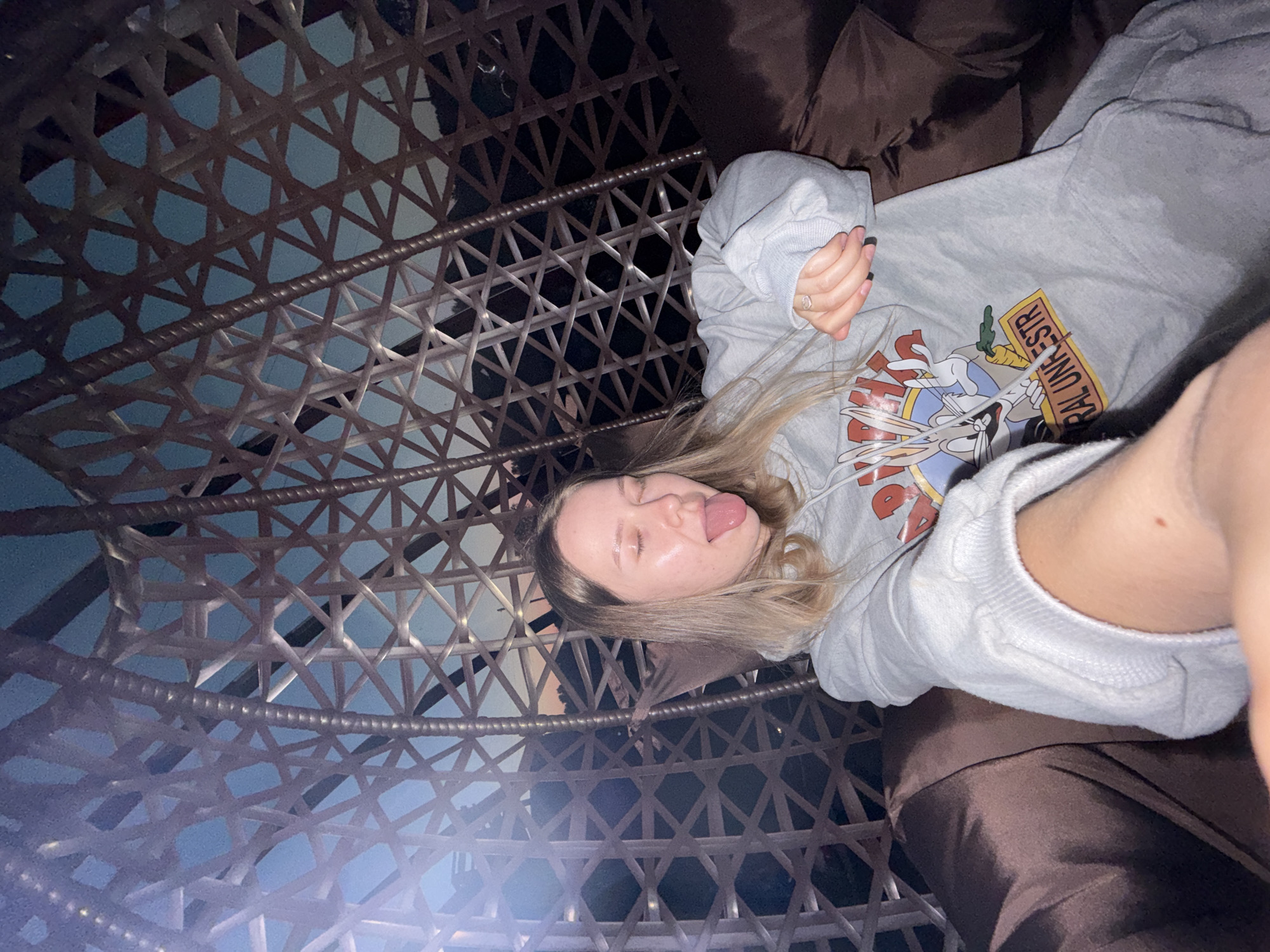 A young, fair-skinned woman with blonde hair lies in what appears to be a hanging wicker chair. She is wearing a gray hooded sweatshirt featuring a cartoon design and white drawstrings. The woman is sticking her tongue out and looking at the camera with her eyes partially closed. The wicker chair has a brown cushion. The background through the wicker is a mix of dark and light areas. There is no information suggesting that this is located in Zhigalki, Russia.