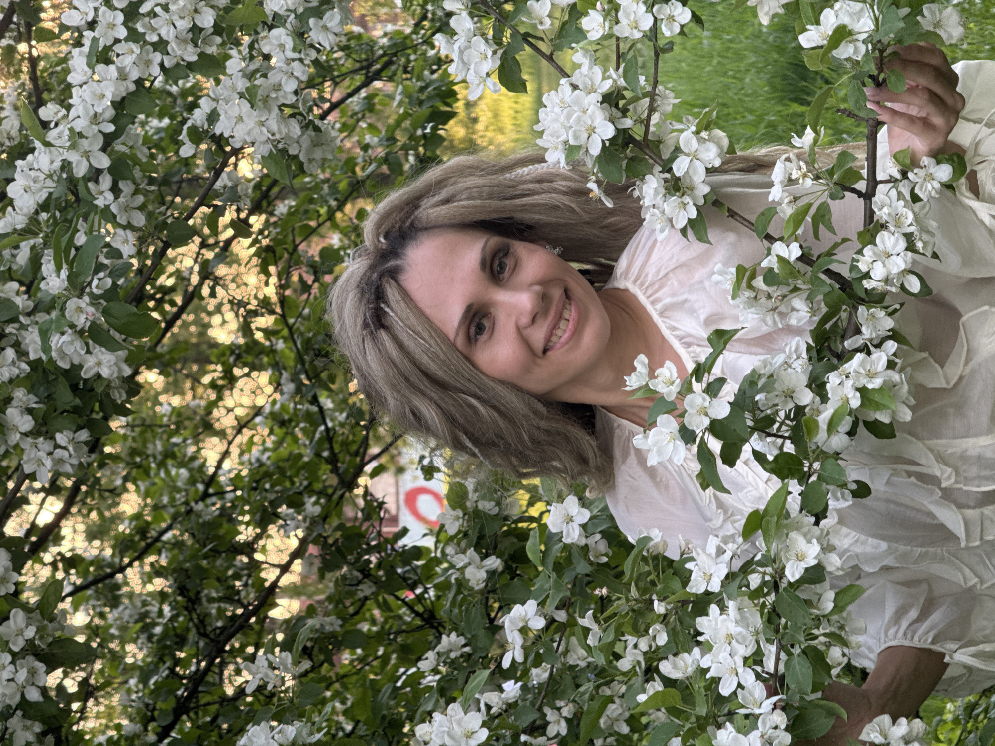 In a daylight outdoor setting, a woman poses within a blooming tree in Шебунино, Russia. The woman, fair-skinned with light-colored hair, smiles directly at the viewer. She wears a light-colored blouse with ruffles. She appears surrounded by the white blossoms of the tree. Her hand is seen gently holding a branch. The tree's foliage fills much of the frame. The background contains hints of greenery and dappled sunlight. A building with a red and white sign is partially visible in the background, obscured by the branches.