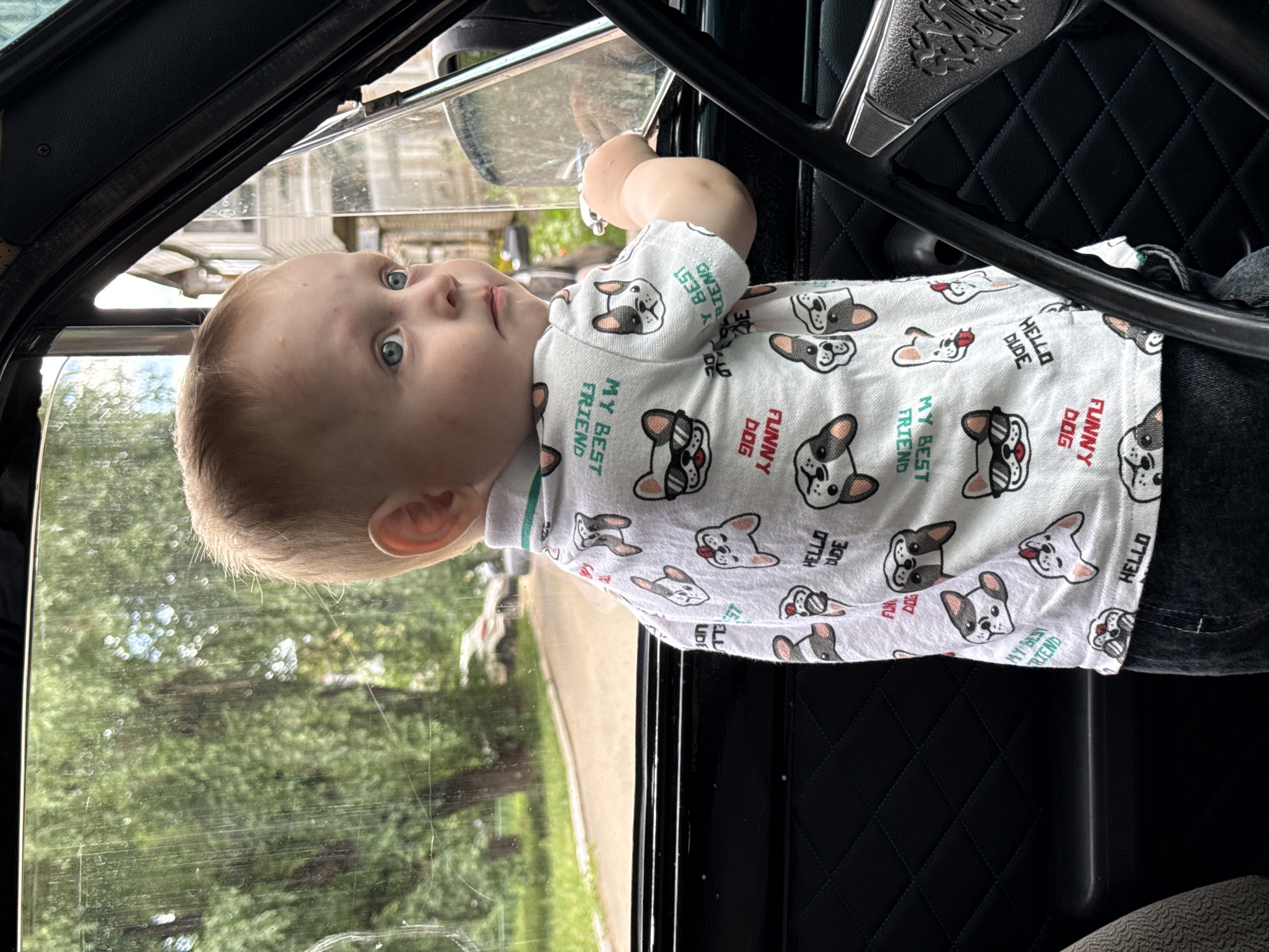 A Caucasian toddler with light hair and blue eyes is sitting in the passenger seat of a vehicle, looking out the window. The child is wearing a white t-shirt with a pattern of French bulldogs and text that reads "MY BEST FRIEND" and "FUNNY DOG". The interior of the car is visible, including a black dashboard and steering wheel. Outside the window, green trees and a street can be seen, suggesting an outdoor, likely urban or suburban setting in the city of Chaikovsky, Russia. The window is slightly smudged.