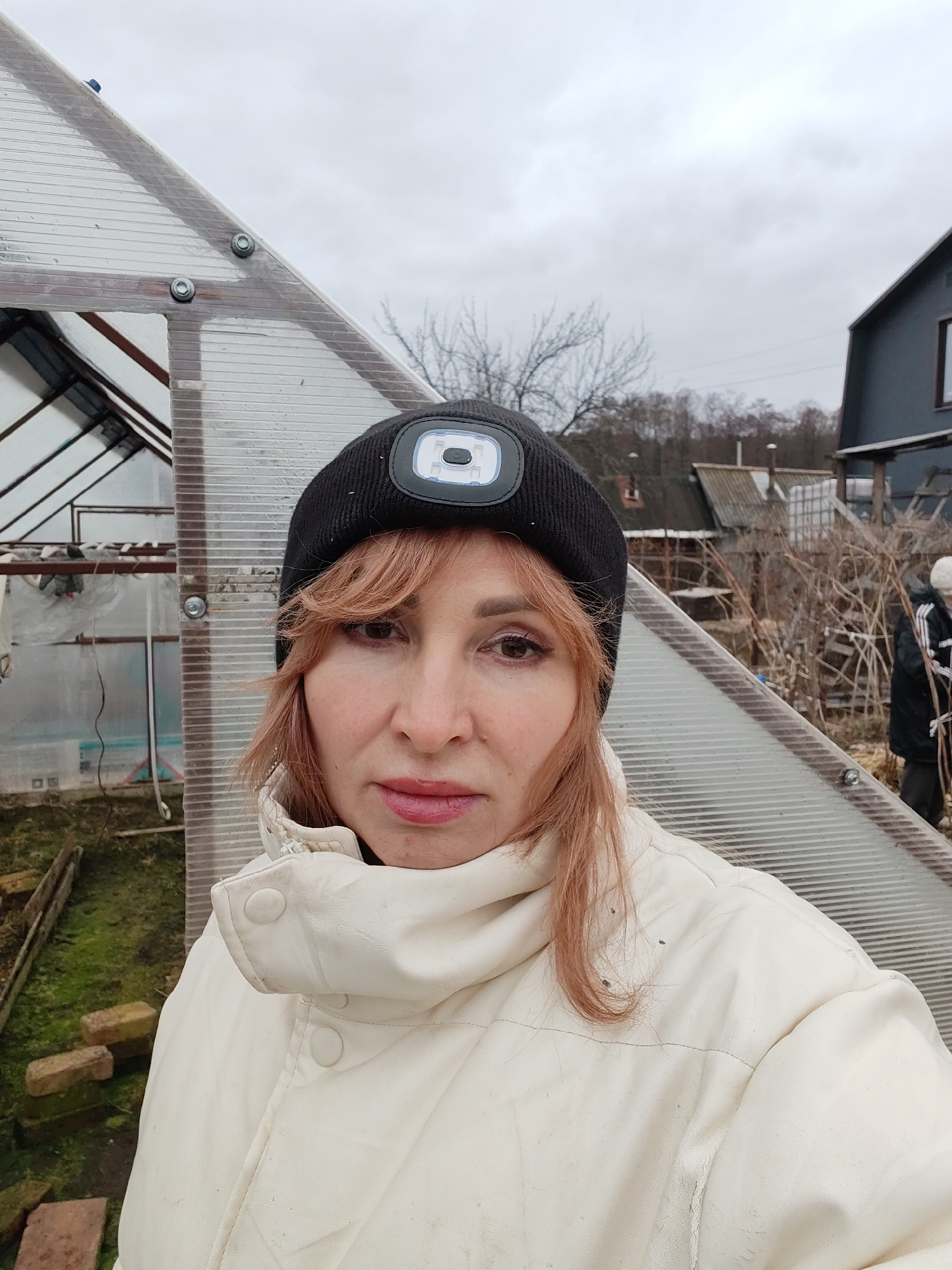 A woman with reddish-brown hair, wearing a cream-colored jacket and a black beanie with an integrated LED light, is the central subject of this outdoor, selfie-style photograph. She has a neutral expression.
Behind her, to the left, stands a greenhouse made of transparent, corrugated polycarbonate panels, bolted together. The interior of the greenhouse shows bare soil and some structural elements. To her right, in the background, another person in a dark jacket with white stripes (possibly an Adidas track jacket) and a light-colored beanie is partially visible, facing away and appearing to be tending to a patch of bare, twiggy bushes or trellises. Further in the background, bare deciduous trees and a dark-colored house are visible. In the lower left foreground, several bricks are scattered on ground covered with green moss or algae.
The sky is overcast and grey, suggesting a cool or cold day, consistent with the subjects' warm clothing. The bare trees and dormant garden elements indicate that the season is likely late autumn, winter, or early spring. The overall setting appears to be a rural or semi-rural garden plot in Chaikovsky, Russia. There is no discernible text visible in the image.