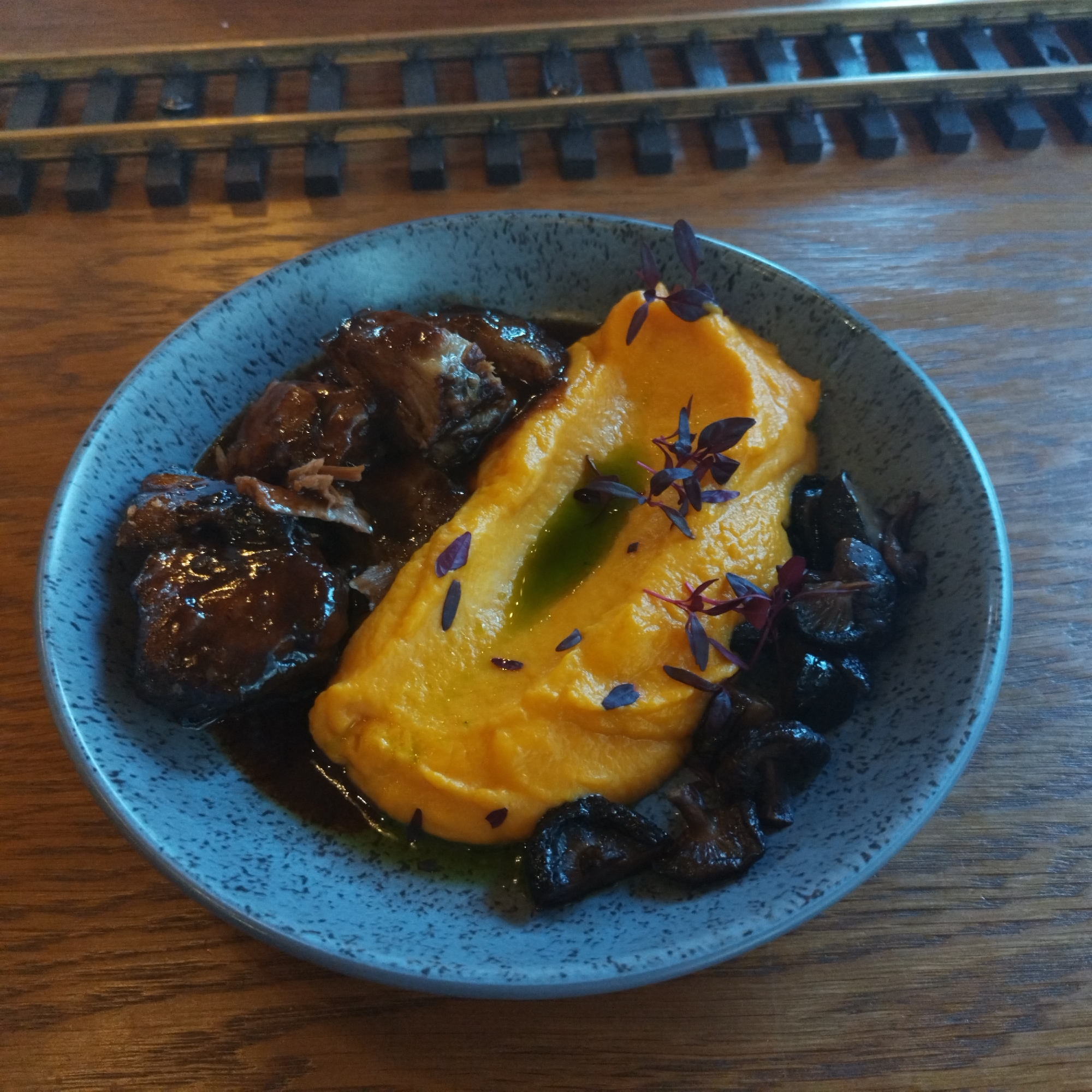 A close-up, overhead shot captures a meticulously plated gourmet meal served in a speckled blue-grey ceramic bowl, resting on a warm-toned wooden table. The dish features three main components: a generous portion of dark, braised meat or stew, glistening with a rich sauce, occupies the left side of the bowl. Centrally placed is a vibrant, smooth orange purée, likely sweet potato or carrot mash, with a drizzle of green oil and scattered small purple sprigs as garnish. Dark, sautéed mushrooms are arranged on the right side of the purée.
In the background, slightly out of focus, a section of a model railway track is visible, complete with metal rails and dark wooden sleepers, indicating a distinct train-themed setting, possibly a restaurant, cafe, or pub. The lighting is soft and warm, consistent with an indoor dining environment. No people are visible, and there is no discernible text. The scene suggests a meal being presented or enjoyed, with attention paid to both the culinary preparation and the unique ambiance of the location.