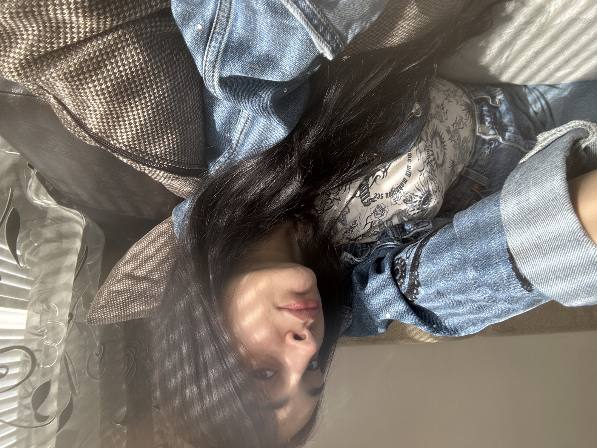 The image depicts a woman with long, dark hair lying on a patterned sofa, possibly indoors. She's wearing a denim jacket over a printed top. Light streams in from the left, possibly through a window with a decorative curtain. There's no information to ascertain the location as the city of Burgas, Bulgaria. The subject's location is indeterminate.