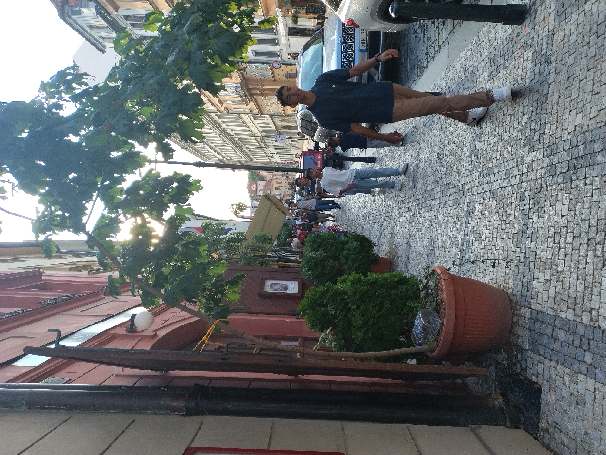 In Prague, Czechia, on a cobblestone street lined with buildings, a young man in a blue polo shirt and brown pants walks towards the camera. He is on the right side of the frame, with other pedestrians on the sidewalk to his left. In the background, several cars are parked, including a silver Jeep. Trees with green leaves hang over the street, and awnings extend from some of the buildings. The sun is setting, casting a warm glow on the scene.
