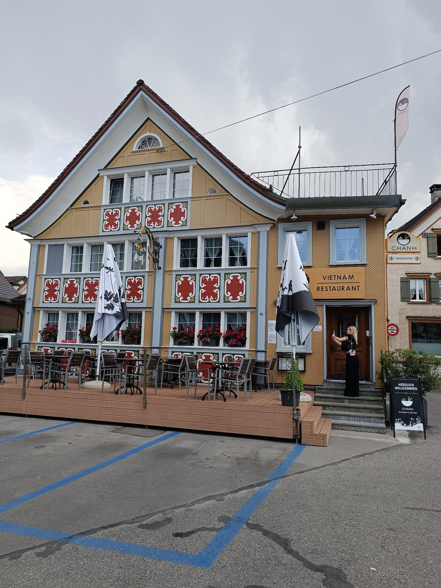 In Appenzell, Switzerland, a two-story building with a yellow facade, adorned with red and white decorative elements, houses the "VIETNAM RESTAURANT." The ground floor features an outdoor seating area with multiple tables and chairs arranged on a wooden deck, protected by umbrellas. A woman in a black dress is standing on the steps leading to the entrance of the restaurant, holding a smartphone and appearing to take a picture. A sign at the entrance reads "HERZLICH WILLKOMMEN" (Welcome) and advertises "CHANH RESTAURANT & TAKE AWAY." The street in front of the restaurant has blue parking lines. The sky is overcast.