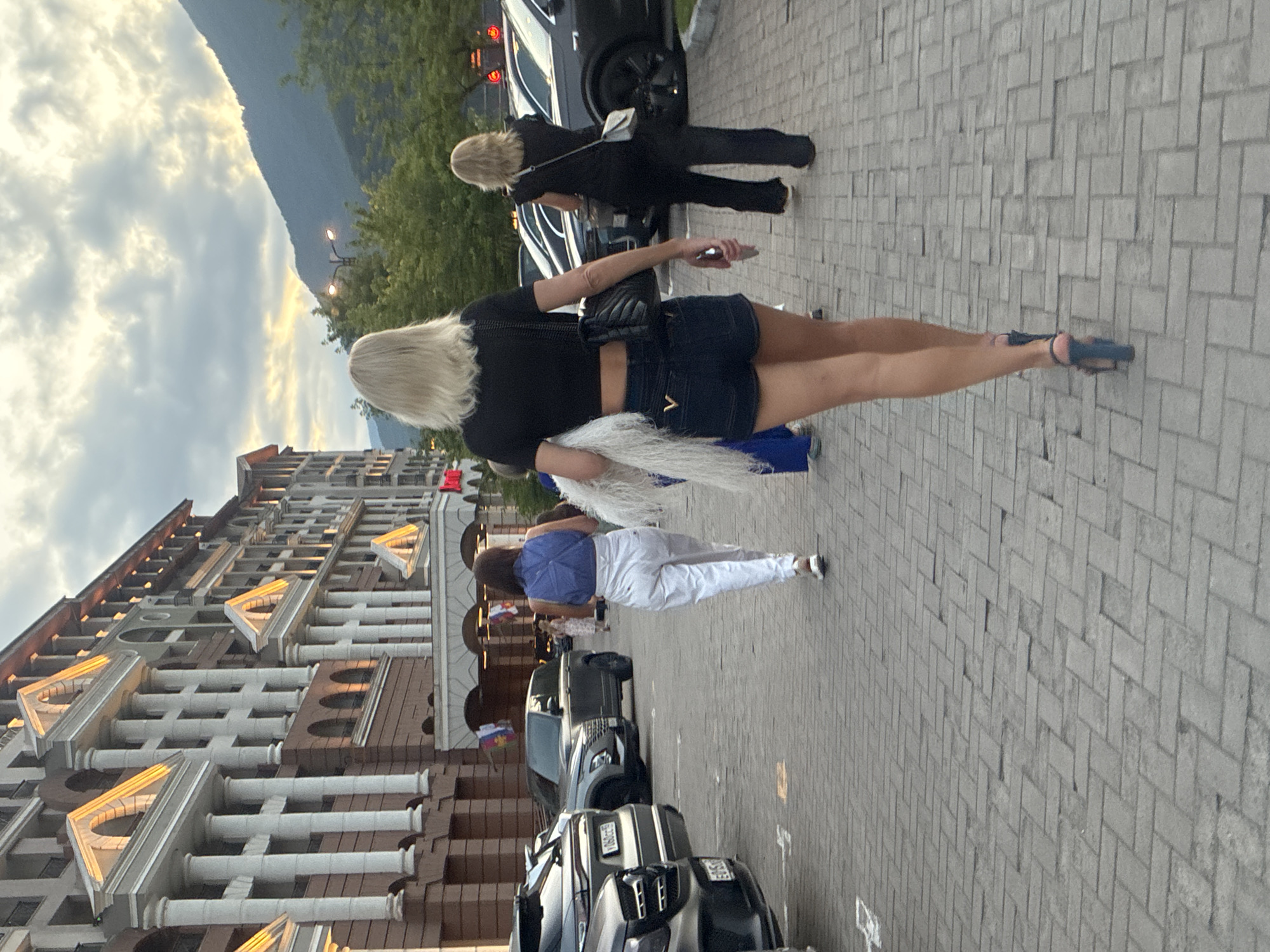 A group of people walk along a cobblestone pathway in front of a grand hotel in Estosadok, Russia. The hotel, with its classical architectural style featuring columns and arched windows, is lit with warm lighting. In the background, lush green trees and a misty mountain range are visible under a cloudy sky. Several cars are parked along the left side of the pathway. The person in the foreground, a woman with blonde hair, walks away from the camera wearing a black crop top, denim shorts, and high-heeled sandals, carrying a white furry accessory. To her left, another woman in white pants and a blue top walks away, and further back, a woman in black pants and a black top also walks away. The scene appears to be captured during the late afternoon or early evening.