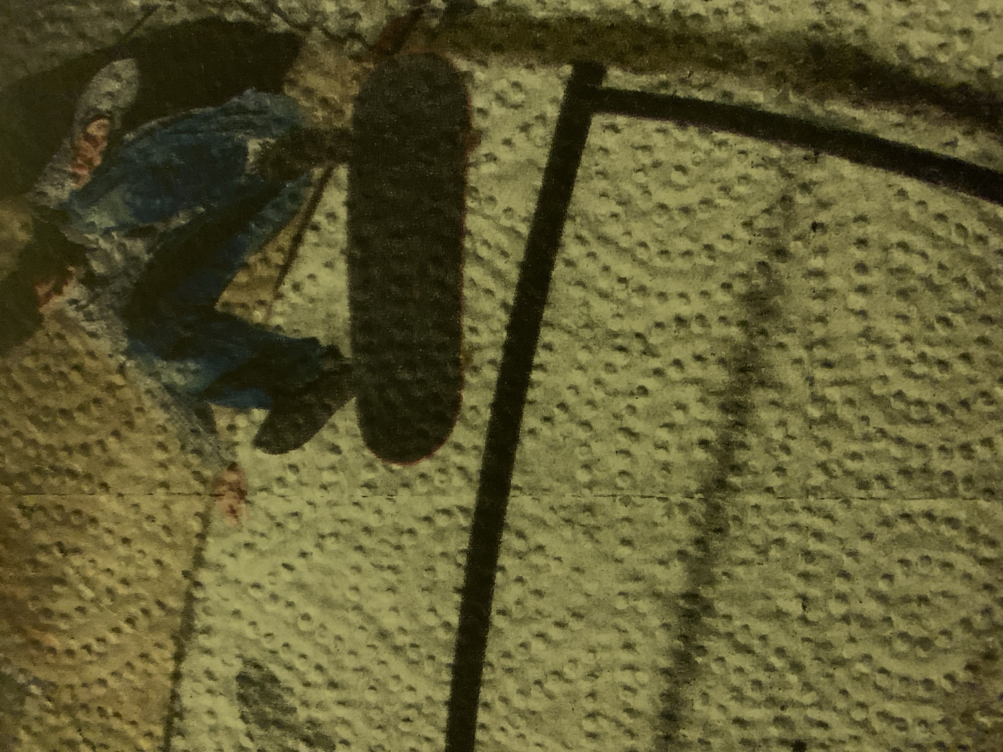 The image shows a person in blue clothing leaning against a black metal structure. The person is partially obscured. A dark, elongated object is positioned near the person and the metal structure. The background has a mottled, textured appearance. The image's overall tonality is muted and indistinct, making it difficult to discern specific details. There is no clear evidence to suggest this image is from the city of Barcelona, Spain.