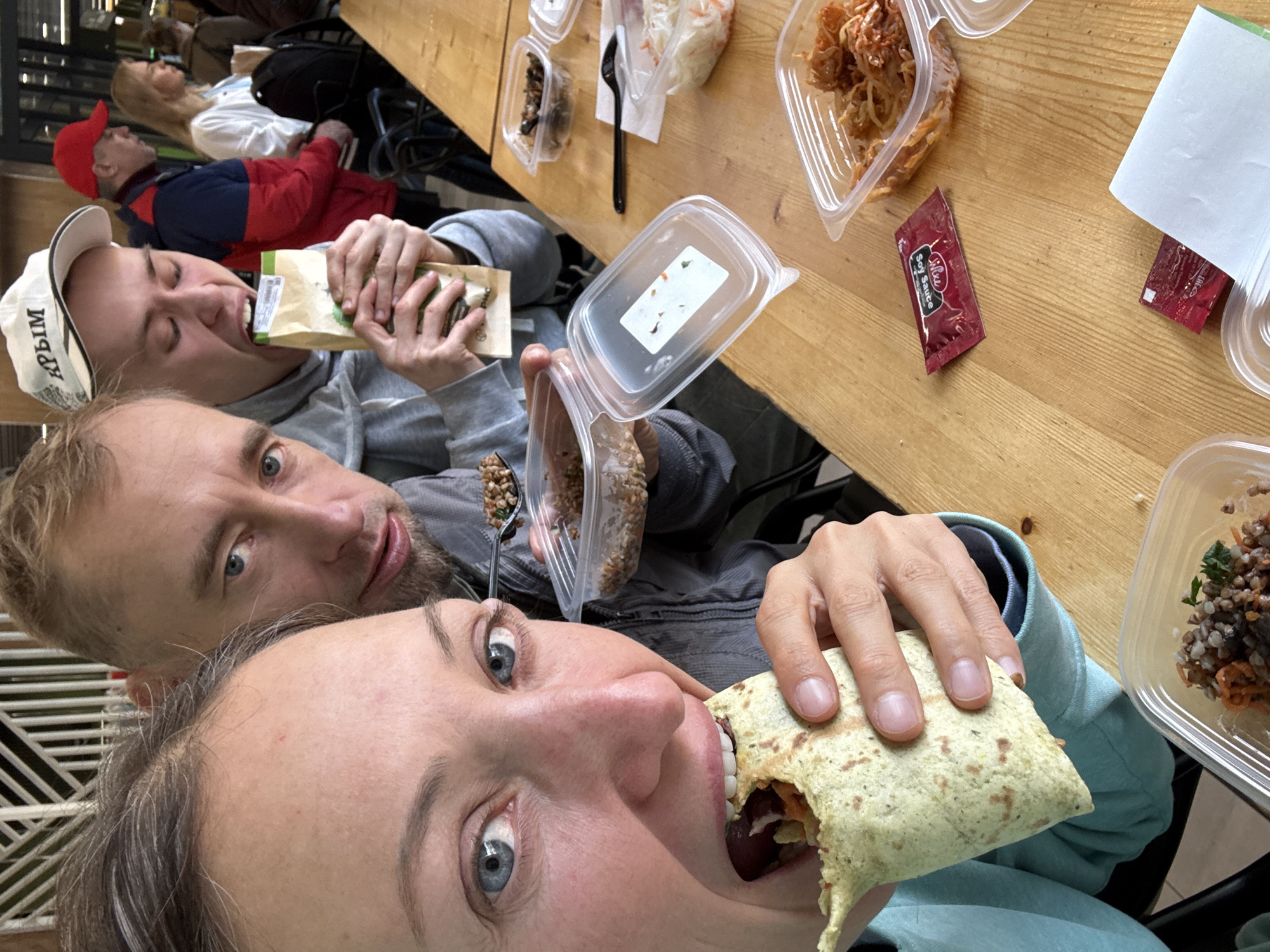 The image captures a candid moment of several people sharing a casual meal at a wooden table in what appears to be an indoor eatery or food court during daytime. In the immediate foreground, a woman in a light blue top is seen with wide, playful eyes looking towards the camera, taking a bite out of a large wrap. Behind her, a man with a beard and a grey hoodie eats from a clear plastic container with a spoon, also looking towards the camera.
Further back, another man wearing a red and blue jacket and a red cap is focused on eating from a paper bag or pouch, his eyes seemingly closed. He also wears a white cap with the Cyrillic text "КРЫМ" (KRYM / Crimea) visible on its brim. The wooden table is laden with various clear plastic containers holding different food items, including grain-based salads, shredded vegetable salads, and a reddish, possibly spicy, dish. A packet of "Wai-Ke Soy Sauce" is discernible on the table alongside another red condiment packet. In the blurred background, other patrons can be seen, contributing to the public dining setting. The scene conveys a relaxed and informal gathering as the group enjoys their meal.