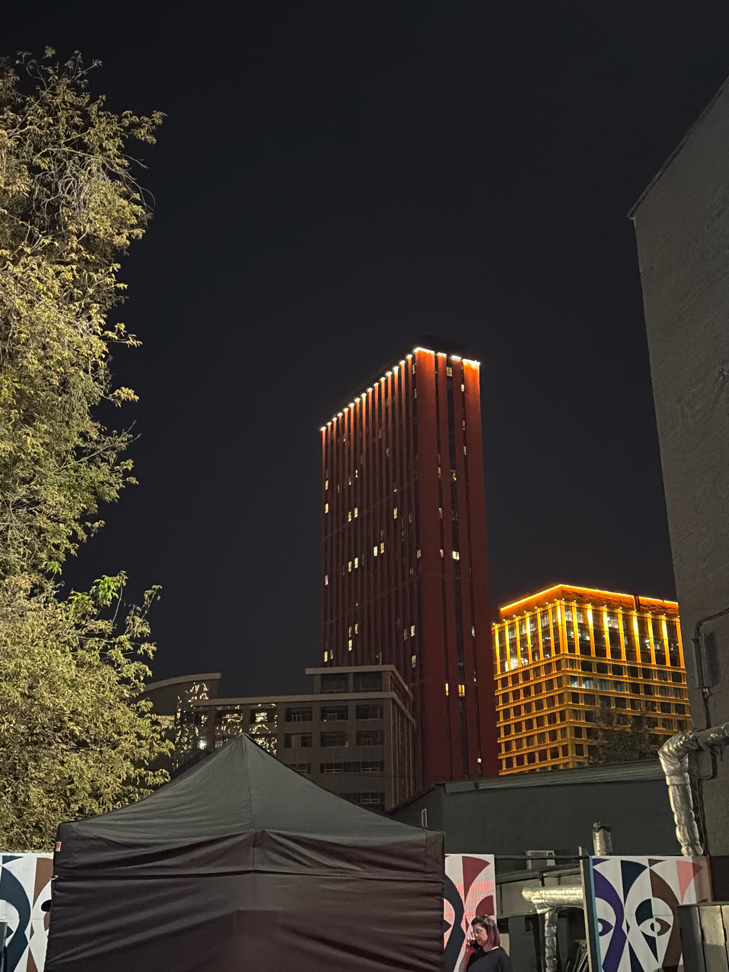 This is a nighttime, eye-level, medium shot of an urban scene in Khimki, Russia. In the foreground, a dark, pitched tent covers the lower portion of the frame. To the left of the tent, a tree with sparse, yellow-green foliage is illuminated by artificial light. Behind the tent, a low building with visible pipes and a mural featuring abstract eye designs is present. In the background, two tall buildings dominate the skyline. The building on the left is a slender, red tower illuminated by lights along its vertical edges and numerous small windows. To its right, a shorter, wider building glows with a bright orange light along its horizontal and vertical structural elements, with visible windows scattered across its facade. The sky is dark, indicating nighttime.