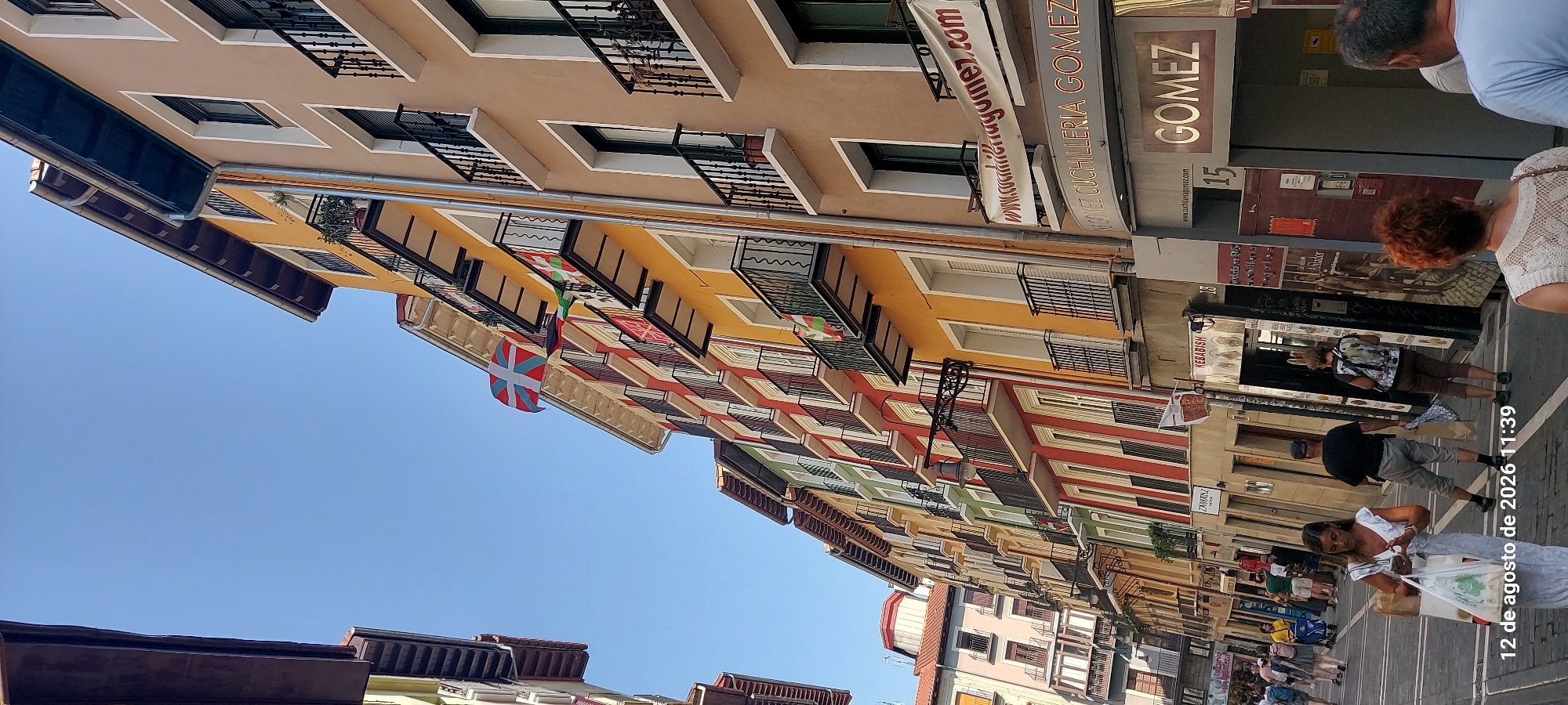 The image depicts a street scene in Pamplona, Spain, likely during daylight hours. The architecture features narrow, colorful buildings with balconies, typical of older European cities. The buildings are multi-story, with many windows and ornate ironwork on the balconies. Flags are visible, including what appears to be the Basque flag, suggesting a possible cultural event or a general display of regional pride.
In the foreground, several people are walking along the street. A woman in a light-colored dress is in the center-left, holding shopping bags and looking at her phone. To her right, a man in a dark t-shirt and shorts walks away from the camera, also carrying a bag. Further back, another woman with a backpack is walking. The street is paved, and there are shop fronts visible along the buildings. One shop sign reads "GOMEZ CUCHILLERIA GOMEZ," indicating a cutlery shop. Another sign indicates "KEBABISH."
The sky is clear and blue, indicating good weather. The perspective is angled upwards, emphasizing the height of the buildings and the narrowness of the street. The timestamp "12 de agosto de 2026 11:39" is overlaid on the bottom left of the image, though the year is in the future. This could indicate a digital overlay or a misinterpretation of the date.