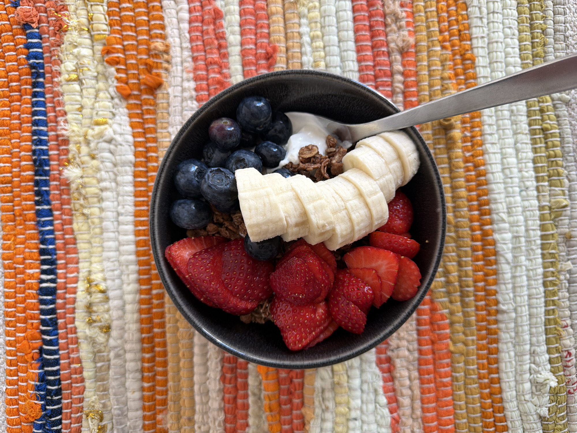 The image depicts a dark-colored bowl filled with a variety of ingredients, presumably for a breakfast or snack meal. The bowl contains sliced strawberries, blueberries, sliced bananas, yogurt, and granola. A silver spoon rests inside the bowl. The bowl sits on a multi-colored woven rug. The location is unknown.