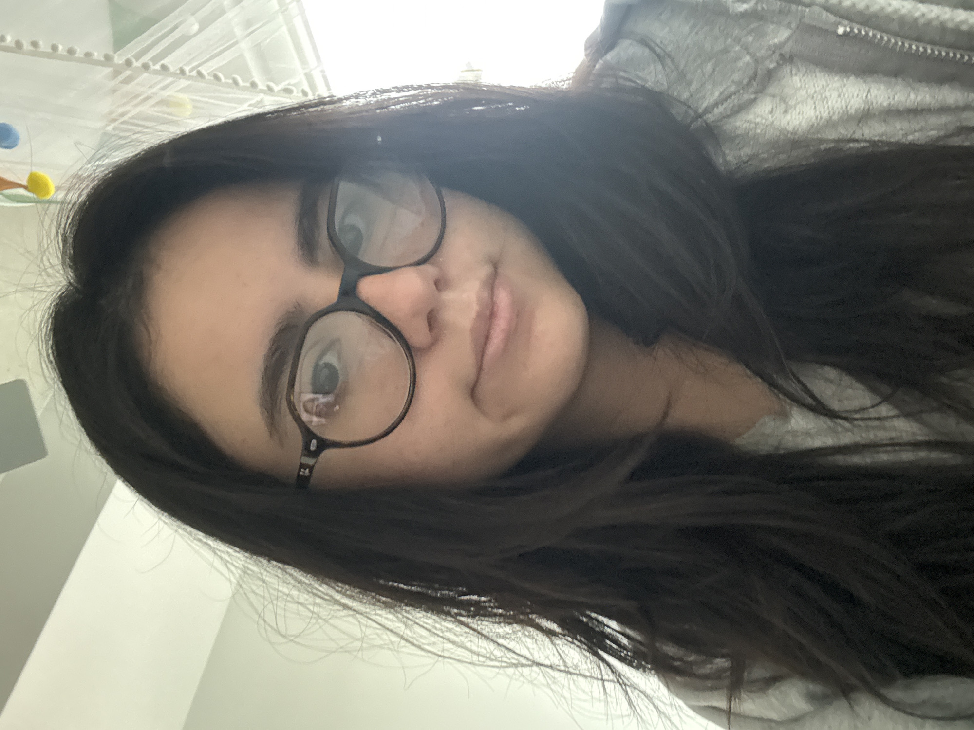 A young woman with long, dark hair and round, black-framed glasses is taking a selfie indoors. She is wearing a gray hooded sweatshirt. The background includes a white wall, a hanging mobile with colorful balls, and part of a ceiling. The location is presumably indoors in an unspecified location within the city of Austin, United States of America.