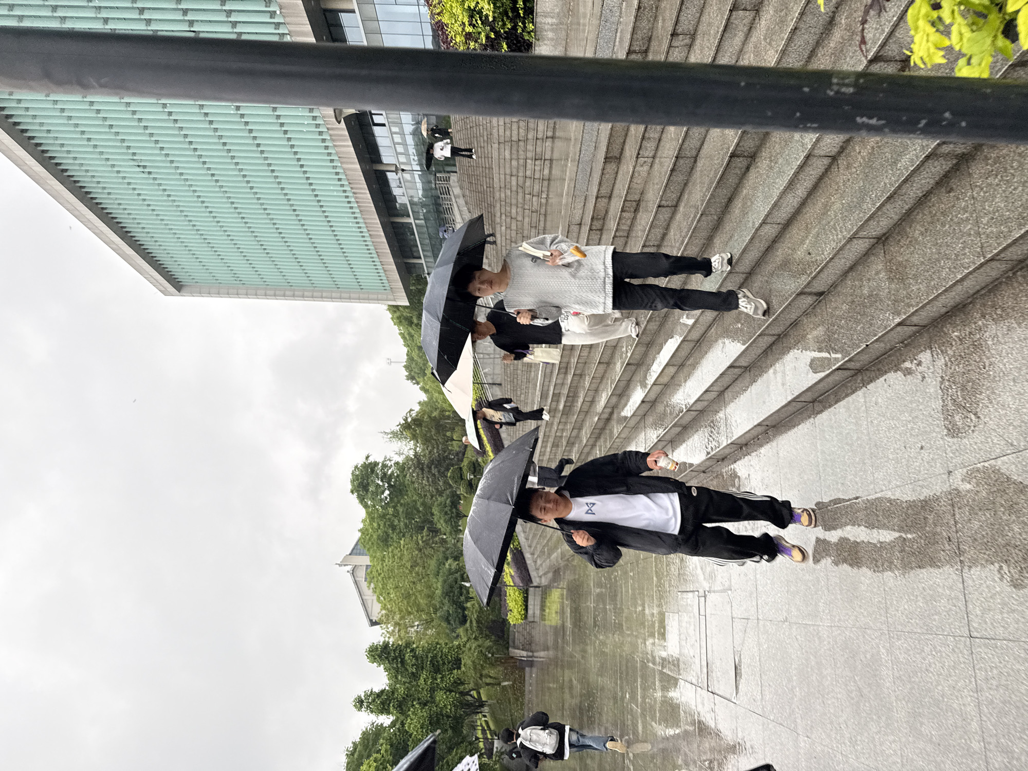 This media file captures an outdoor scene on a rainy, overcast day in Jiangjin, China. The image is oriented vertically (rotated 90 degrees clockwise).
The scene features several individuals walking on wet stone stairs and a paved ground area. Most people are holding black umbrellas, indicating ongoing rainfall. The ground is visibly wet and reflective, showing distorted reflections of people and their umbrellas.
In the foreground, a person walks on the paved area, holding an open black umbrella in one hand and a white bottle in the other. They are wearing a black jacket over a white t-shirt that has a dark "ΣV" or "EV" like emblem, black track pants with white stripes, and dark sandals with purple accents. Further back, on a wide set of stone stairs, another person in a light grey sweater and black pants is walking, holding a black umbrella, an open book, and a snack. Behind them, two more individuals are ascending the stairs, also with umbrellas. In the far background, additional people are visible on the stairs, some with umbrellas.
The setting includes a modern building with a distinctive light green or blue-tinted horizontal glass panel structure, possibly an overhang or roof, extending over the stairs. Lush green trees and foliage are visible in the background, contrasting with the grey sky and wet urban environment. The sky is completely overcast, suggesting a dull daytime atmosphere. No specific emotions are discernible on the faces of the subjects, who appear focused on navigating the wet conditions. There is no other visible text apart from the logo on the t-shirt.