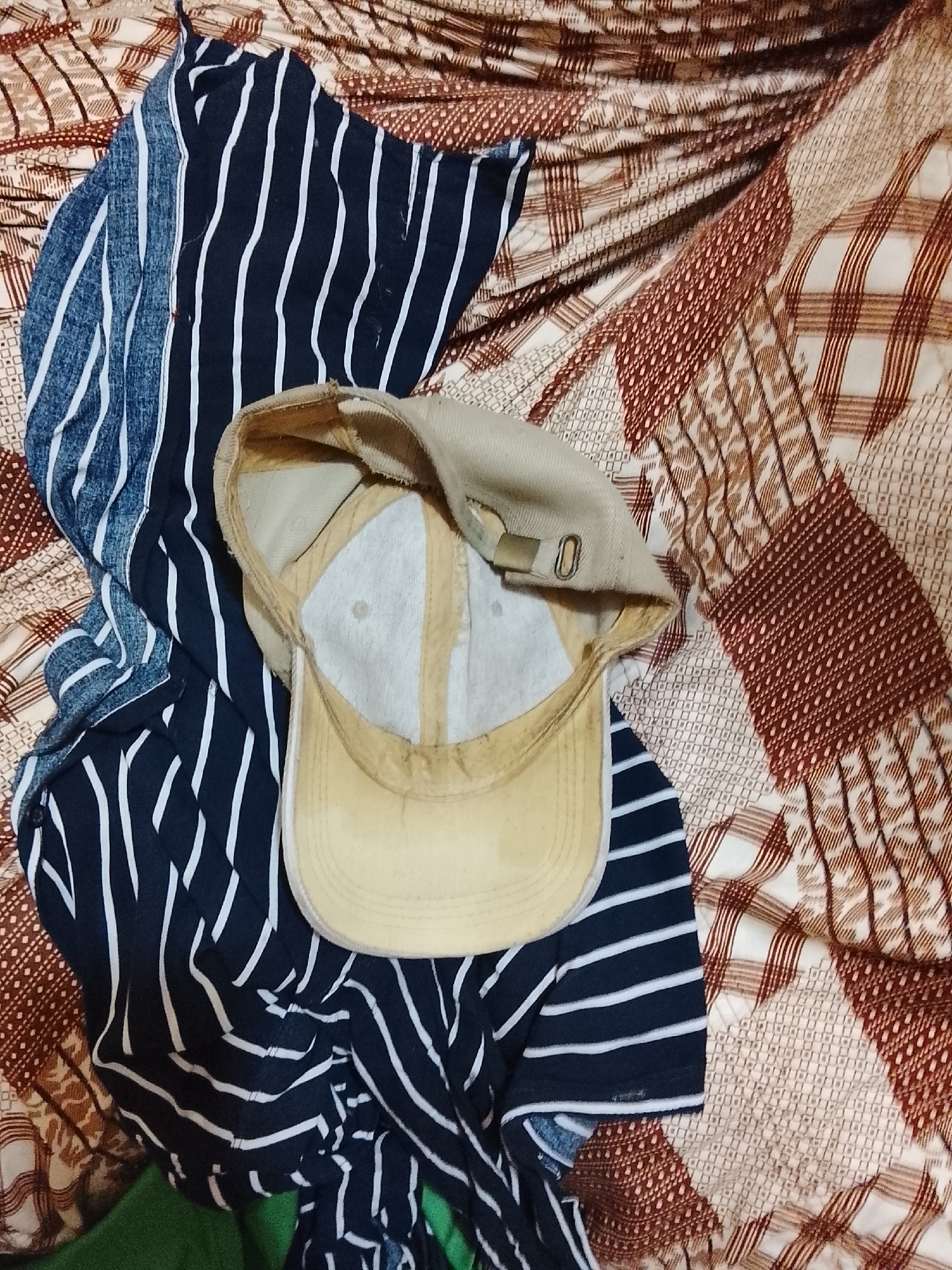 The image shows a tan baseball cap, seen from the inside, resting on a striped navy and white shirt. The shirt is laid out on a patterned beige and brown fabric. The setting appears to be indoors. There is no indication of location or people in the image.