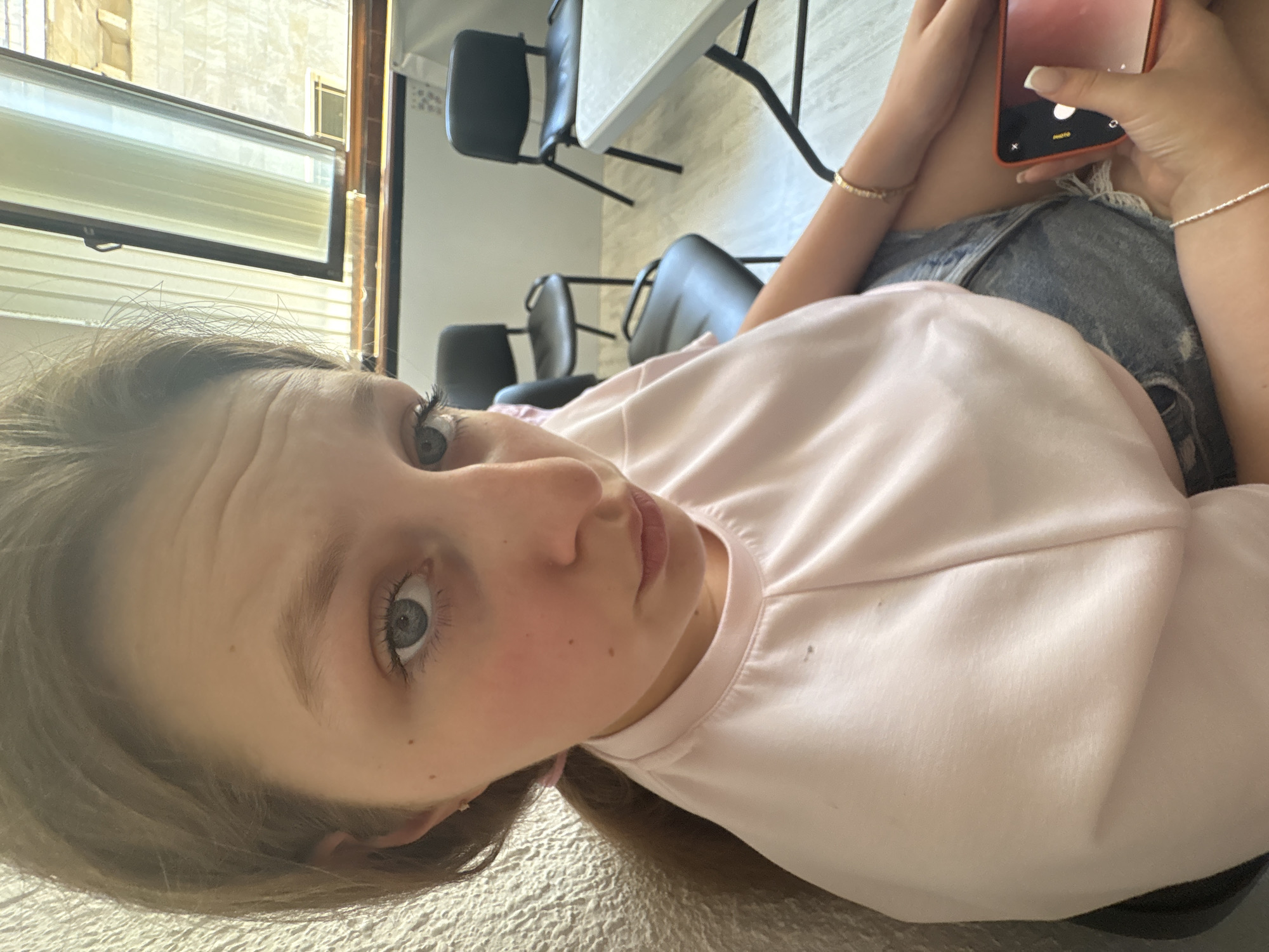 The image shows a young woman sitting indoors. She has light brown hair pulled back into a ponytail and is wearing a light pink t-shirt and denim shorts. She is holding a red smartphone in her right hand. In the background, there are several black chairs and a table, suggesting a classroom or meeting room. The room has a large window with blinds. The overall lighting suggests it is daytime. The background architecture visible through the window appears to be typical of buildings in Burgas, Bulgaria, with light-colored facades and tiled roofs, common in older European cities.