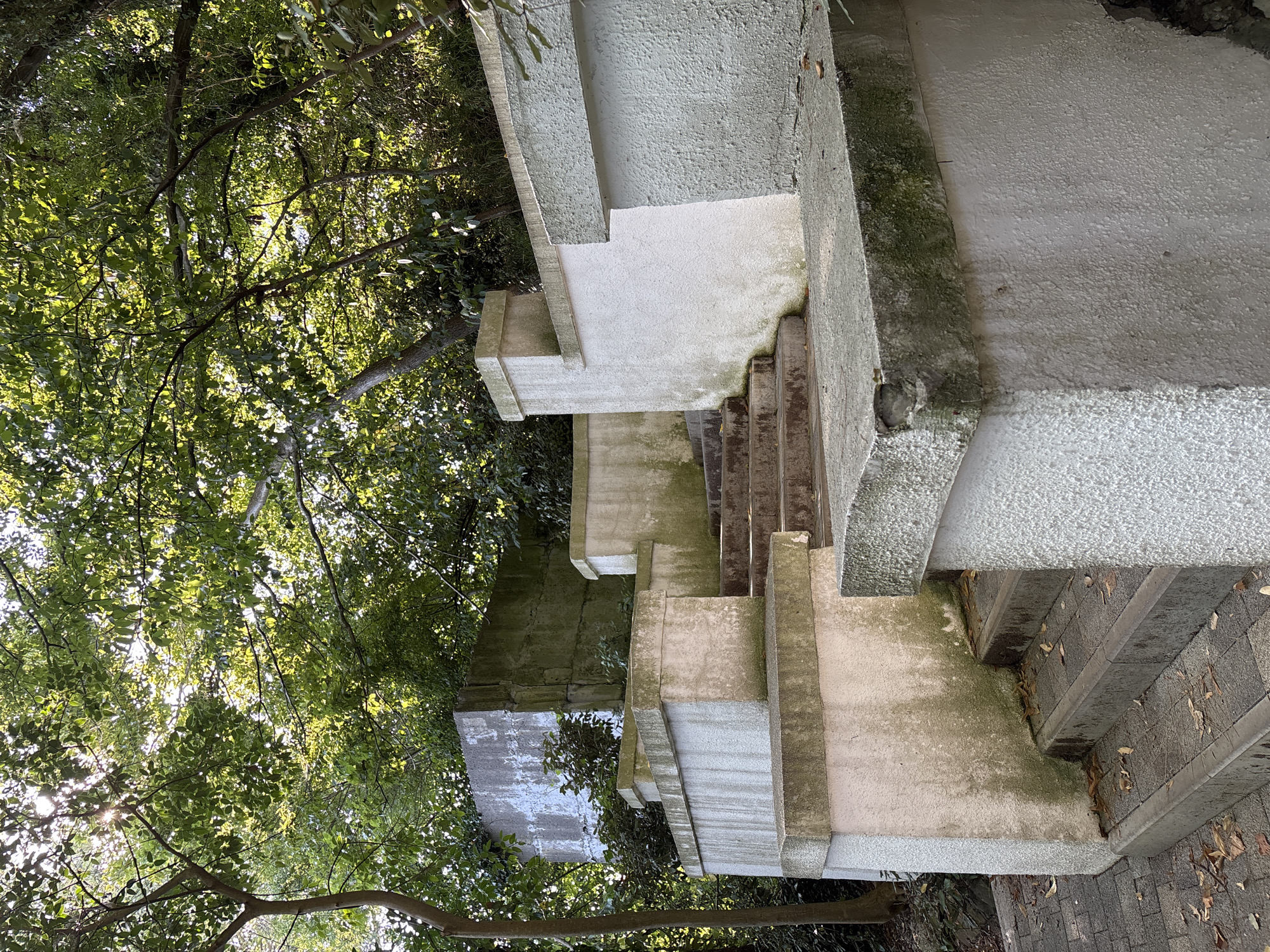 This image depicts a tiered outdoor staircase with white stucco walls and concrete steps, weathered and showing signs of moss and dirt accumulation. The staircase is surrounded by lush green trees, with sunlight filtering through the leaves, creating dappled light. In the background, there's a large, grey concrete structure. The scene appears to be a public outdoor space. There are no people or animals visible in the image.