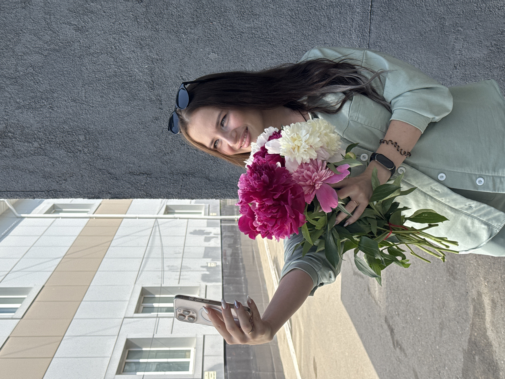 A young woman with long, dark hair and sunglasses perched on her head stands in front of a textured gray wall. She is holding a bouquet of peonies in shades of pink and white, and taking a selfie with her phone, which is in her left hand. She is wearing a light green collared shirt and pants. The background shows a modern building with horizontal beige and white stripes, and windows. The ground is paved and appears to be a street or sidewalk. The overall scene suggests an outdoor, urban environment, possibly in Chaikovsky, Russia, given the architectural style.