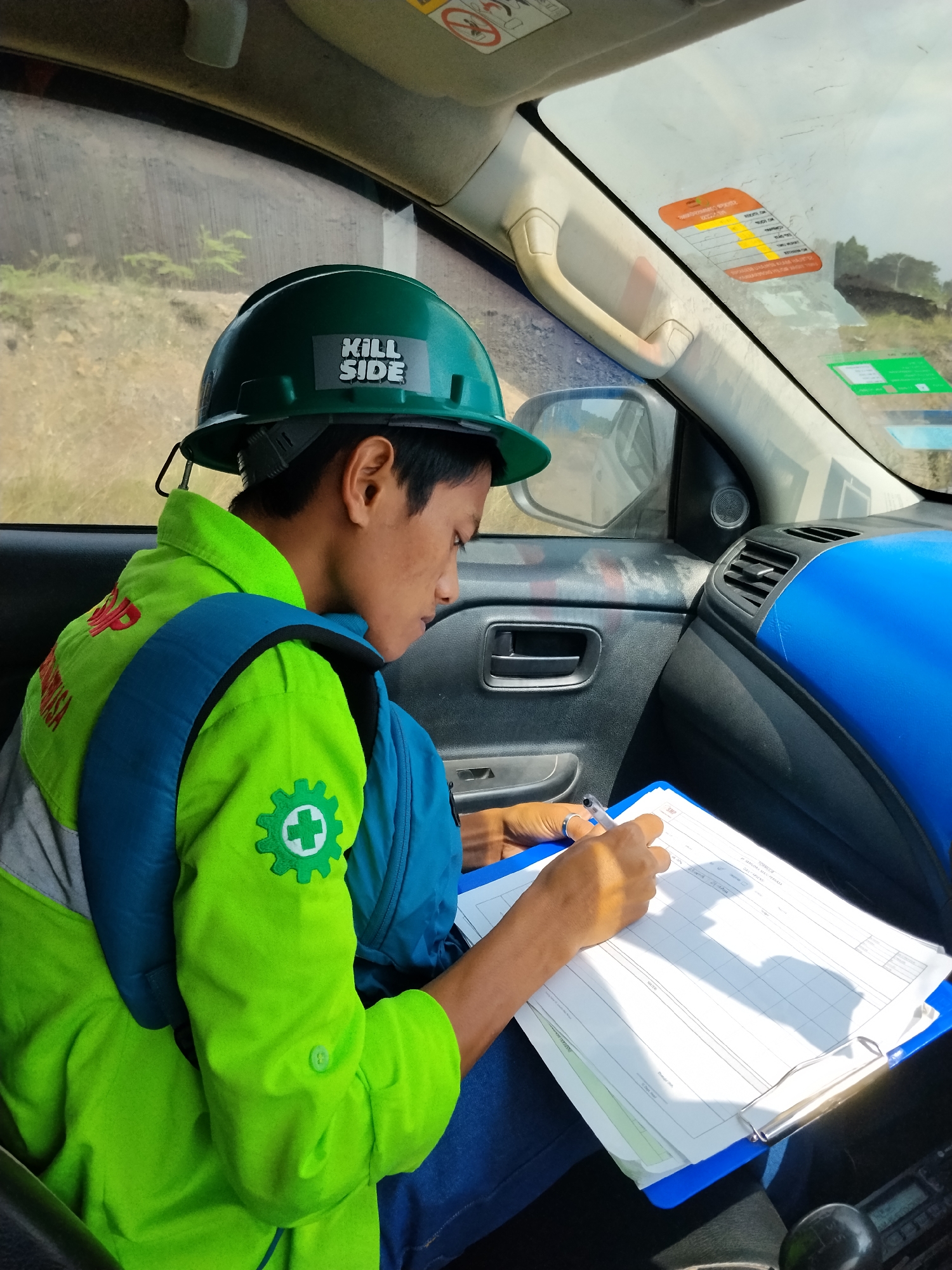A young person wearing a green hard hat with a "KILL SIDE" sticker and a bright green safety vest over a blue backpack is sitting in the passenger seat of a vehicle and writing on a clipboard. The hard hat has a black strap hanging down the back. The safety vest has a white gear emblem with a white cross in the center on the left sleeve, and red text on the back. The person is wearing blue jeans. The vehicle's interior is visible, including the dashboard, a side mirror reflecting an outdoor scene, and a passenger-side door handle. The exterior scene through the windows shows a dry, hilly landscape with some sparse vegetation. Several stickers are visible on the vehicle's windshield, including an orange sticker with text and a green sticker. The location is identified as Sempayau, Indonesia.