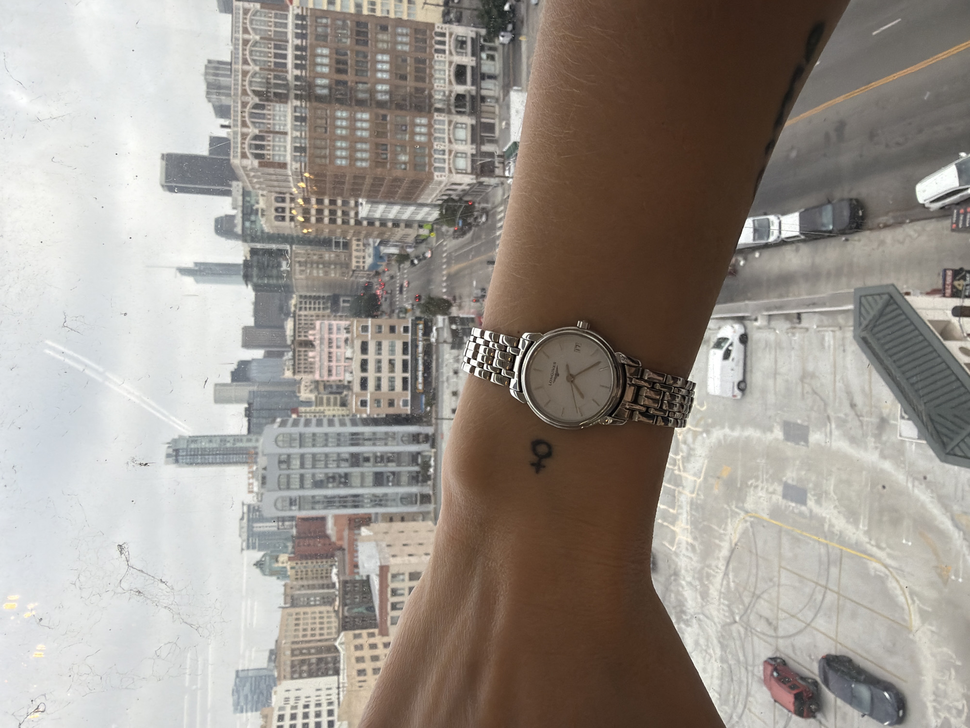 The image shows a person's left forearm with a silver wristwatch and a small tattoo of the Venus symbol on their wrist. The forearm is positioned in the foreground, with a blurred cityscape of Los Angeles, California, in the background. The city skyline features numerous buildings of varying heights and architectural styles, indicative of a downtown urban environment. Streets with cars are visible below the buildings, suggesting a busy city. The sky is overcast. The vantage point appears to be from a window, as evidenced by faint smudges and reflections on a glass surface.