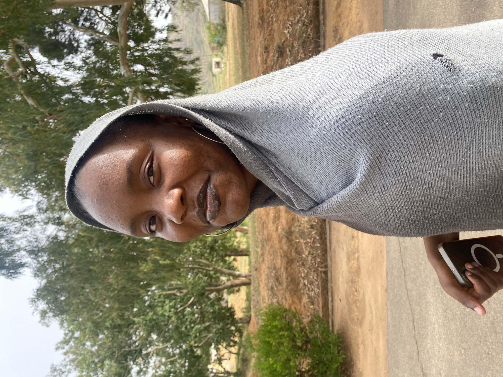 A head-and-shoulders portrait depicts a young Black woman outdoors, likely in Bukuru, Nigeria, during daylight hours. The image is taken from a slightly low angle, looking upwards towards the woman and the trees in the background.
The woman has dark skin, brown eyes, and prominent lips, appearing to have a slight smirk or neutral expression as she looks directly at the viewer. She is wearing a light grey, knitted hoodie or scarf that covers her head and part of her shoulders. A small tear is visible on the upper left side of the fabric. A gold-colored hoop earring can be seen on her right ear.
In her right hand, which is held loosely by her side, she grips a dark-colored smartphone. The phone's case features a distinctive white circular design with a vertical line, resembling a 'P' or a stylized power symbol.
The background consists of an outdoor scene dominated by various trees with green foliage, suggesting a natural or semi-urban environment. The ground appears to be a mix of dry earth and a light-colored concrete path. No specific buildings or structures are clearly discernible, but some distant, blurred elements might indicate sparse construction. The lighting is bright and even, consistent with a clear day. No visible text is identifiable in the image.